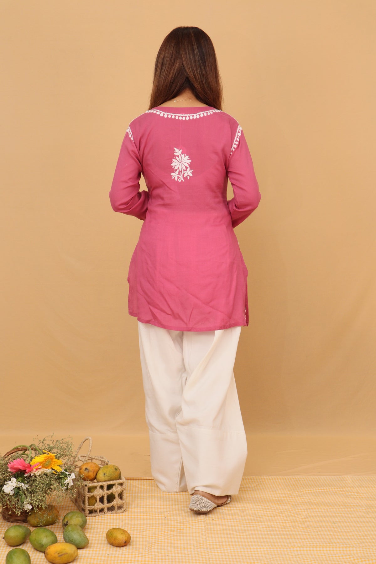 A woman stands with her back to the camera wearing a pink long-sleeve kurta with white embroidery on the back and white wide-leg pants, set against a solid beige backdrop with decorative fruits and flowers on the floor.