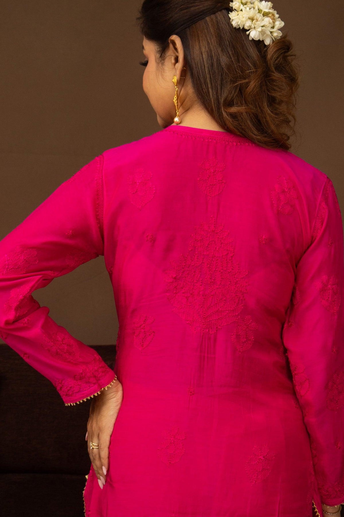 A woman shown from the back wearing a bright fuchsia pink embroidered kurta with long sleeves and delicate gold-toned jewelry against a warm brown background. Pink Muslin Chikankari Bakhiya 2 Piece Set with Golden Gotta Lace