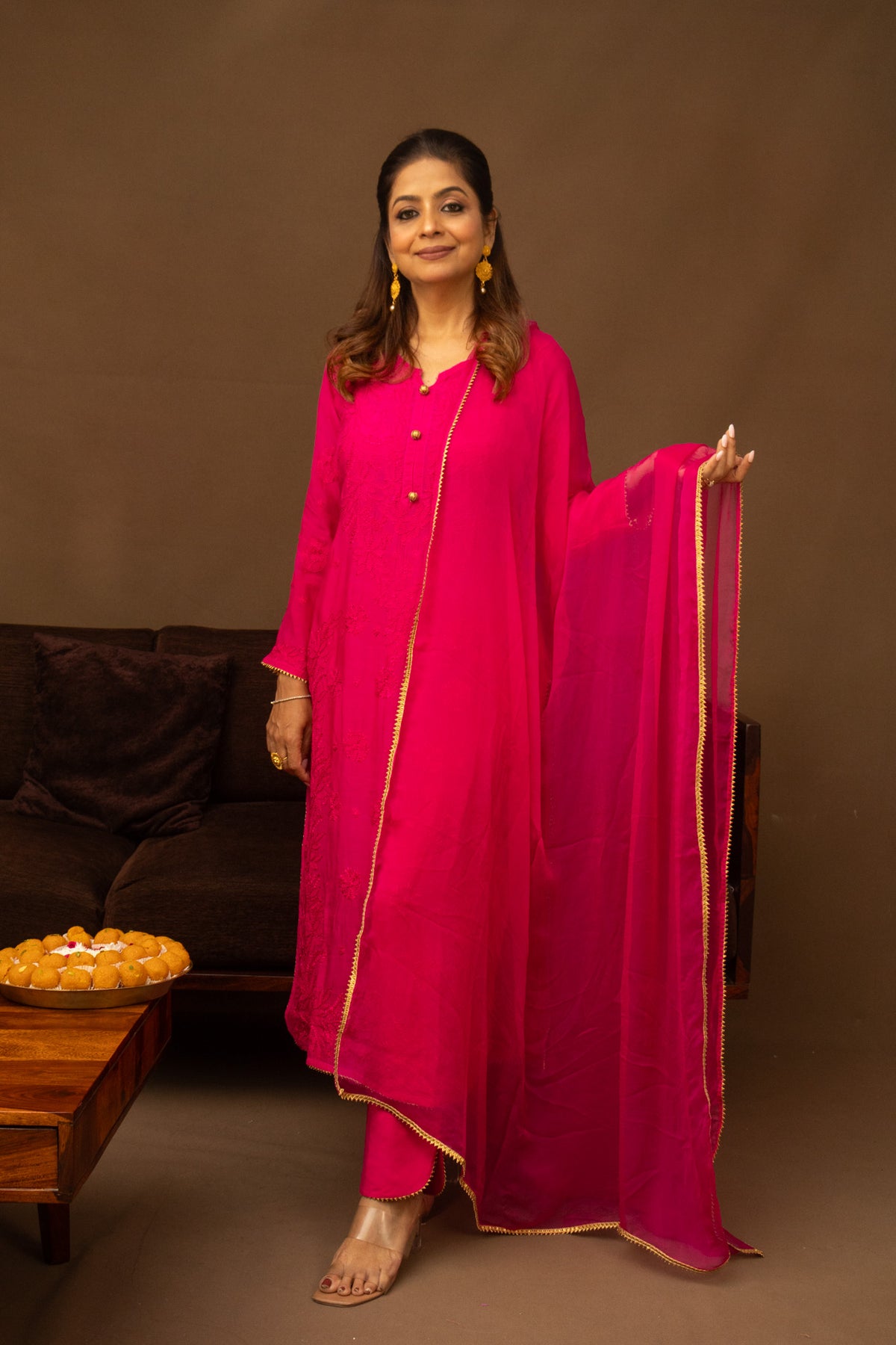 A woman modeling a bright pink traditional outfit with a matching dupatta, featuring gold border accents, set against a warm brown studio background with a sofa and coffee table.