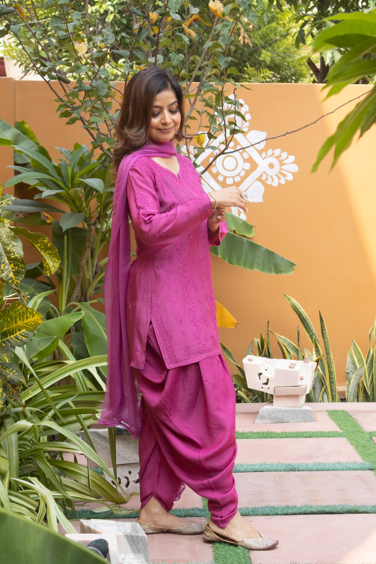 A woman wearing a magenta pink traditional Indian salwar kameez suit with matching dupatta, standing in a garden setting with plants and an orange wall in the background.