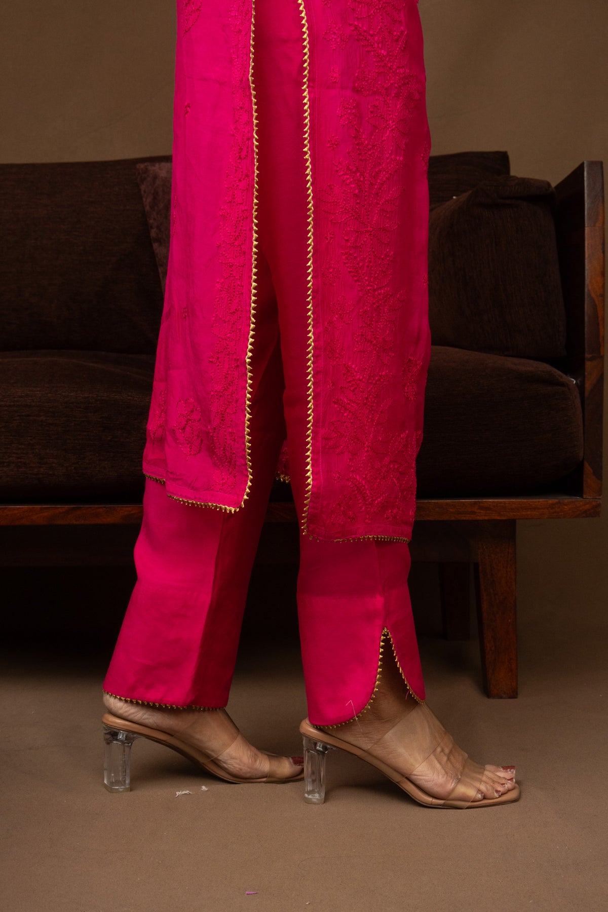 A person wearing bright fuchsia pink traditional pants with gold trim and matching sheer draped fabric, paired with nude heeled sandals, standing in front of a dark brown sofa.