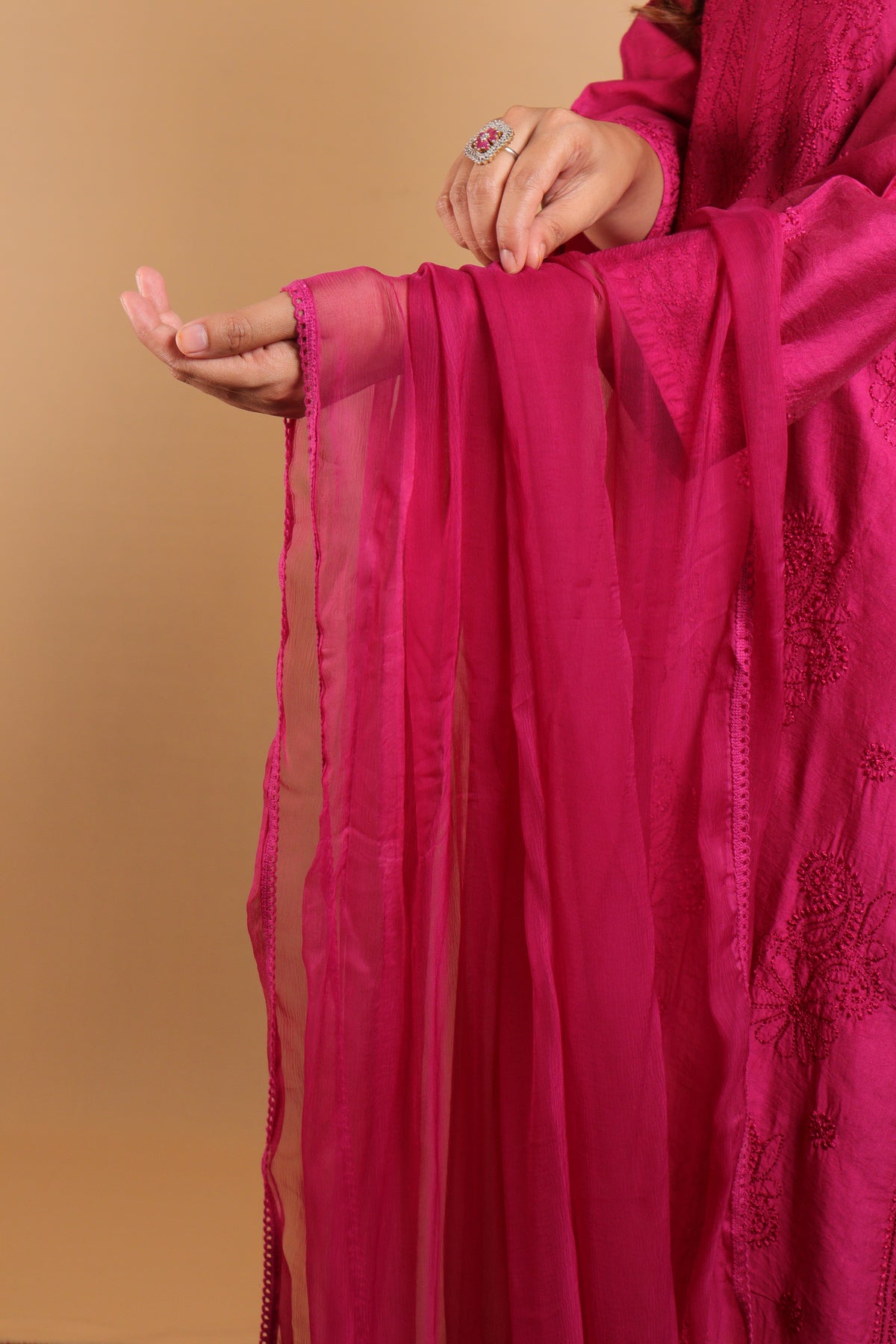 A close-up of a person wearing a bright magenta pink traditional outfit and holding a matching sheer dupatta against a solid beige background.