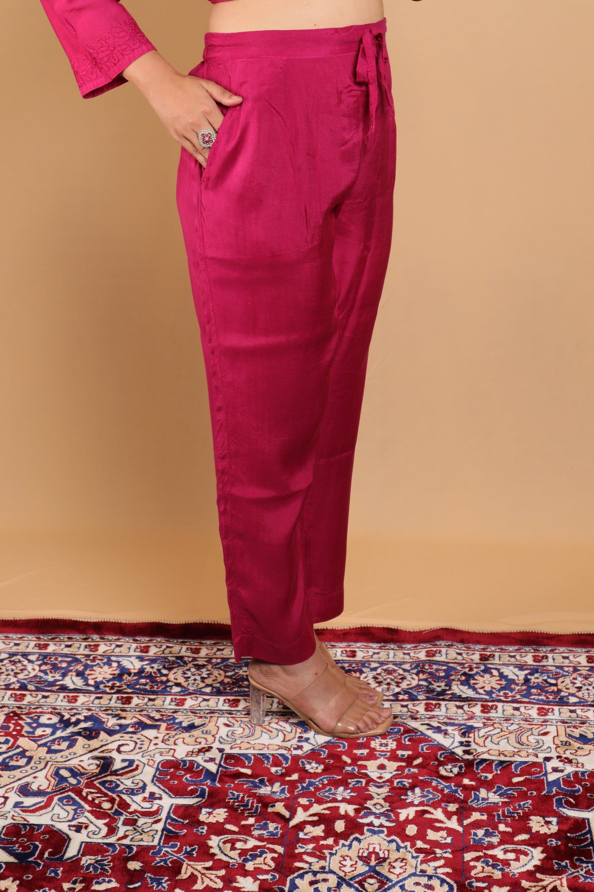 A person wearing bright magenta drawstring trousers and a matching top, standing on a red patterned rug against a beige wall.