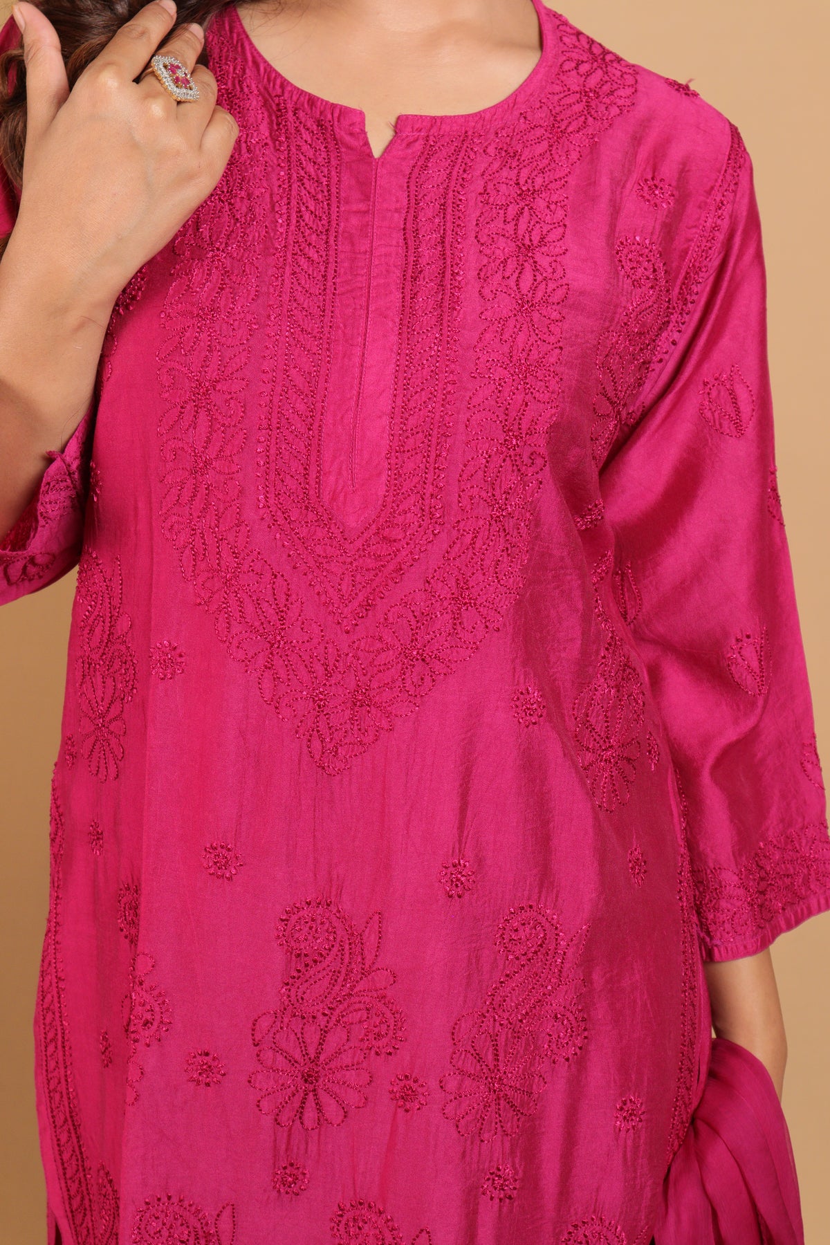 A close-up of a woman wearing a bright pink embroidered kurta with floral and paisley lace detailing, set against a solid beige background.