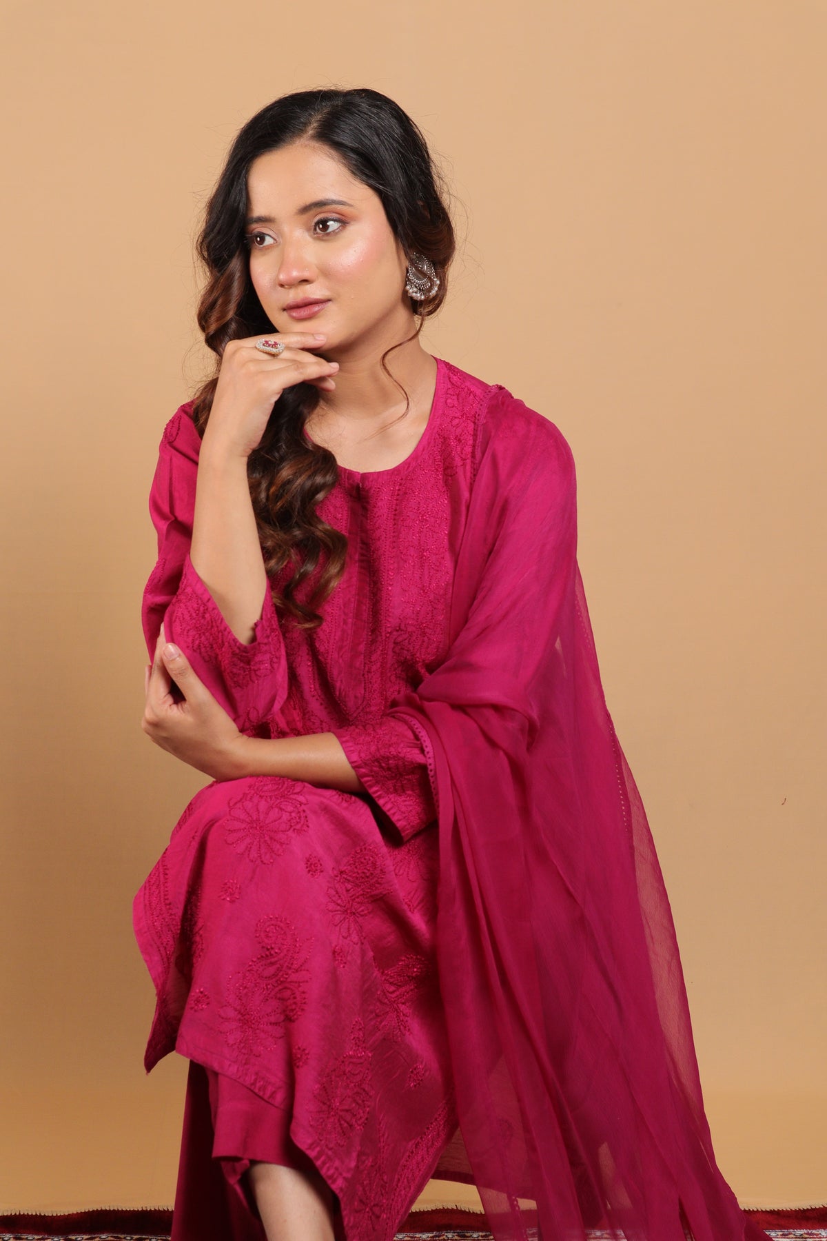 A woman wearing a magenta pink embroidered kurta set with a matching sheer dupatta, posed against a solid beige studio background.