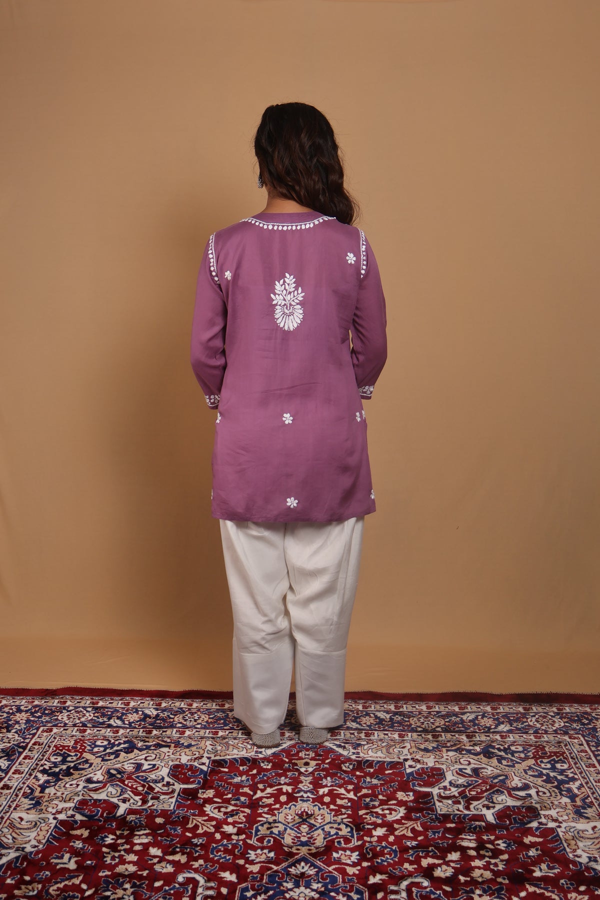 A woman stands with her back to the camera wearing a mauve purple long-sleeve kurta with white embroidery and white pants, standing on a red patterned rug against a solid tan backdrop.
