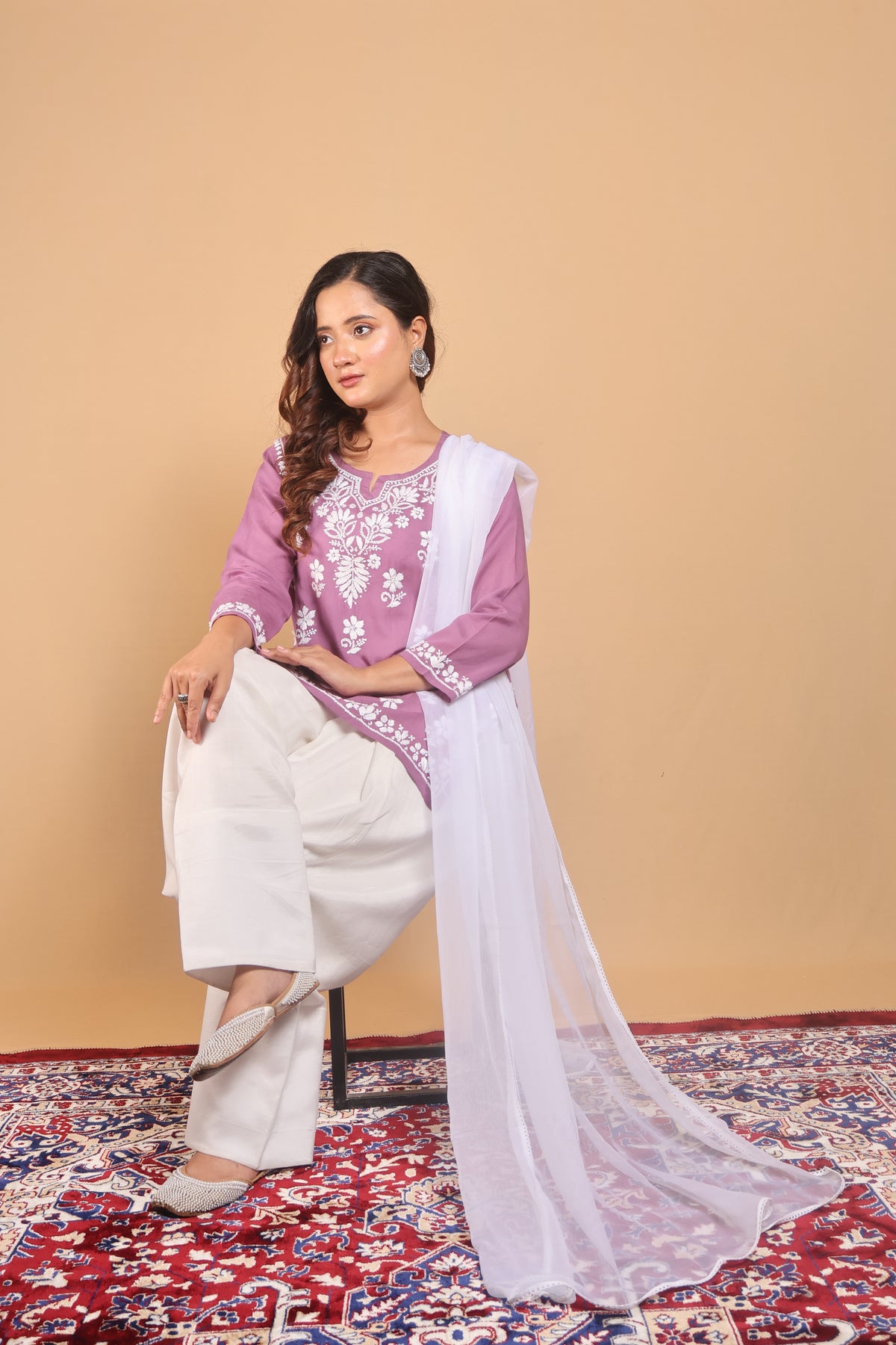 A woman models a traditional Indian outfit consisting of a lavender embroidered kurta, white pants, and a white dupatta, seated on a patterned rug against a solid beige background.