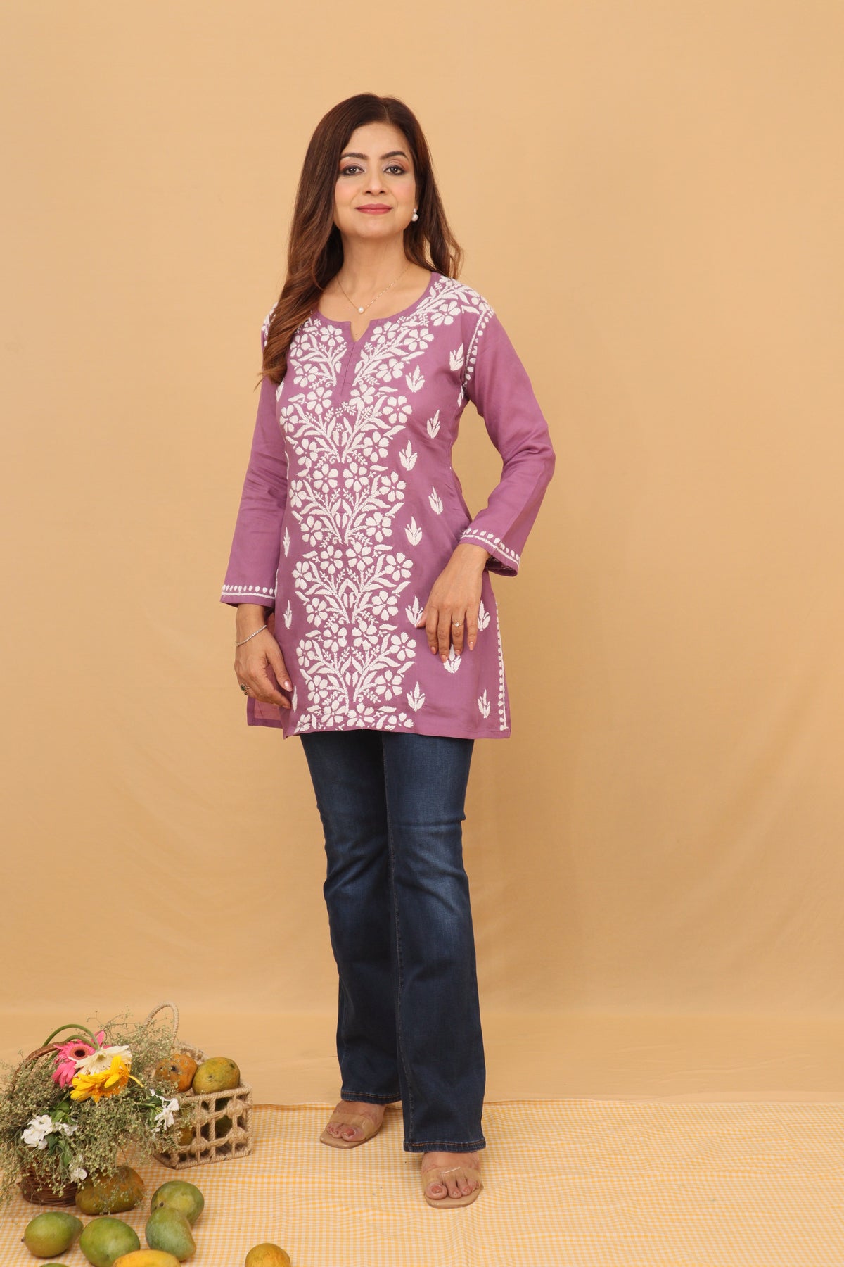 A woman modeling a purple long-sleeve kurta with white floral embroidery, paired with dark blue jeans, standing against a solid beige backdrop.