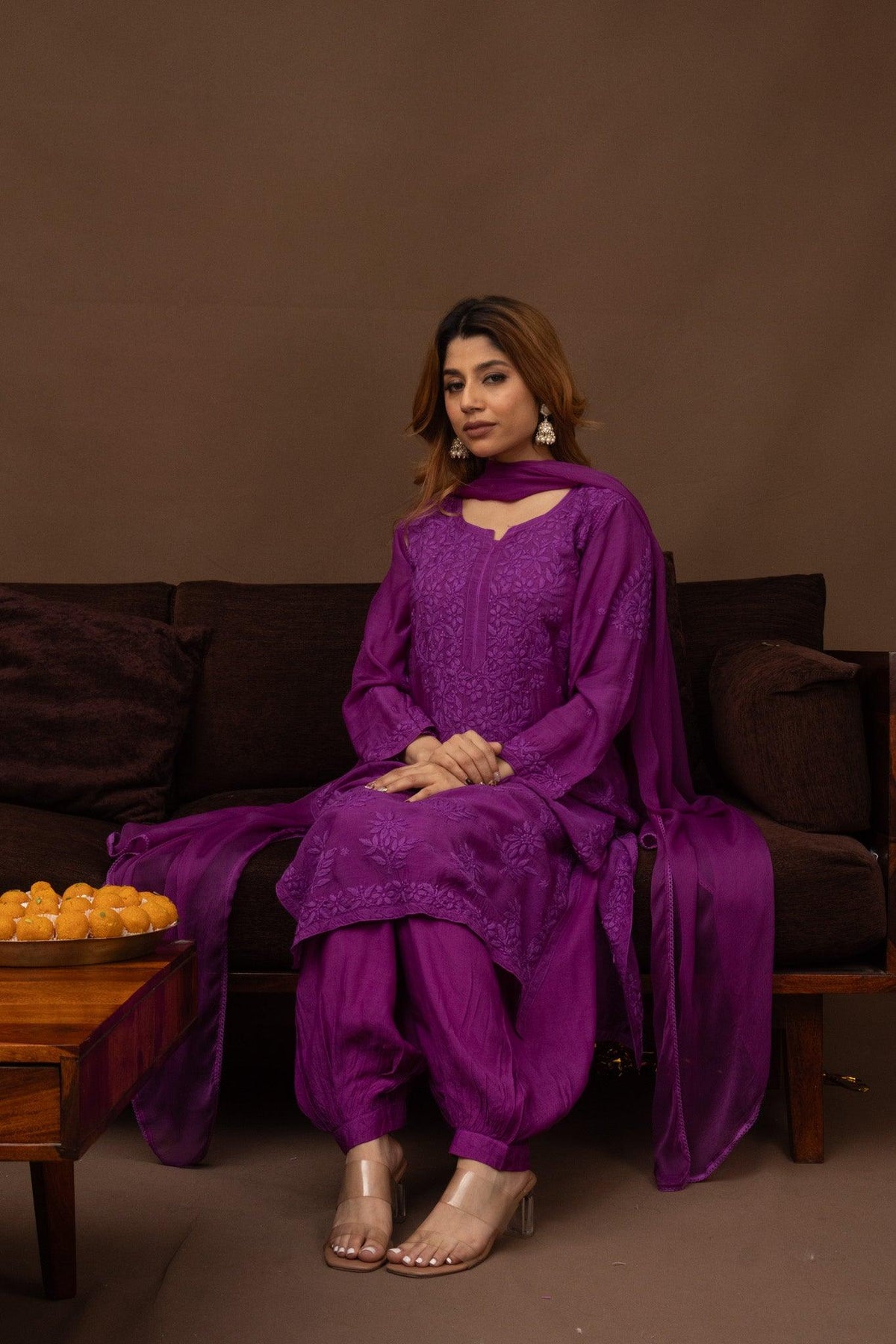 A woman models a traditional purple salwar kameez suit with matching dupatta while sitting on a dark brown couch. The outfit features intricate embroidery on the kameez top and matching purple trousers. Purple Muslin Chikankari Ghaaspatti Kurti with Afghani Pants 3 Piece Set
