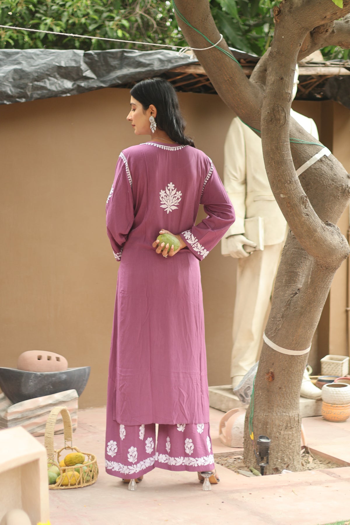 A woman stands outdoors wearing a long magenta-purple traditional kurta with white floral embroidery on the back and sleeves, paired with matching embroidered palazzo pants. She is photographed from behind near a tree and a beige wall.