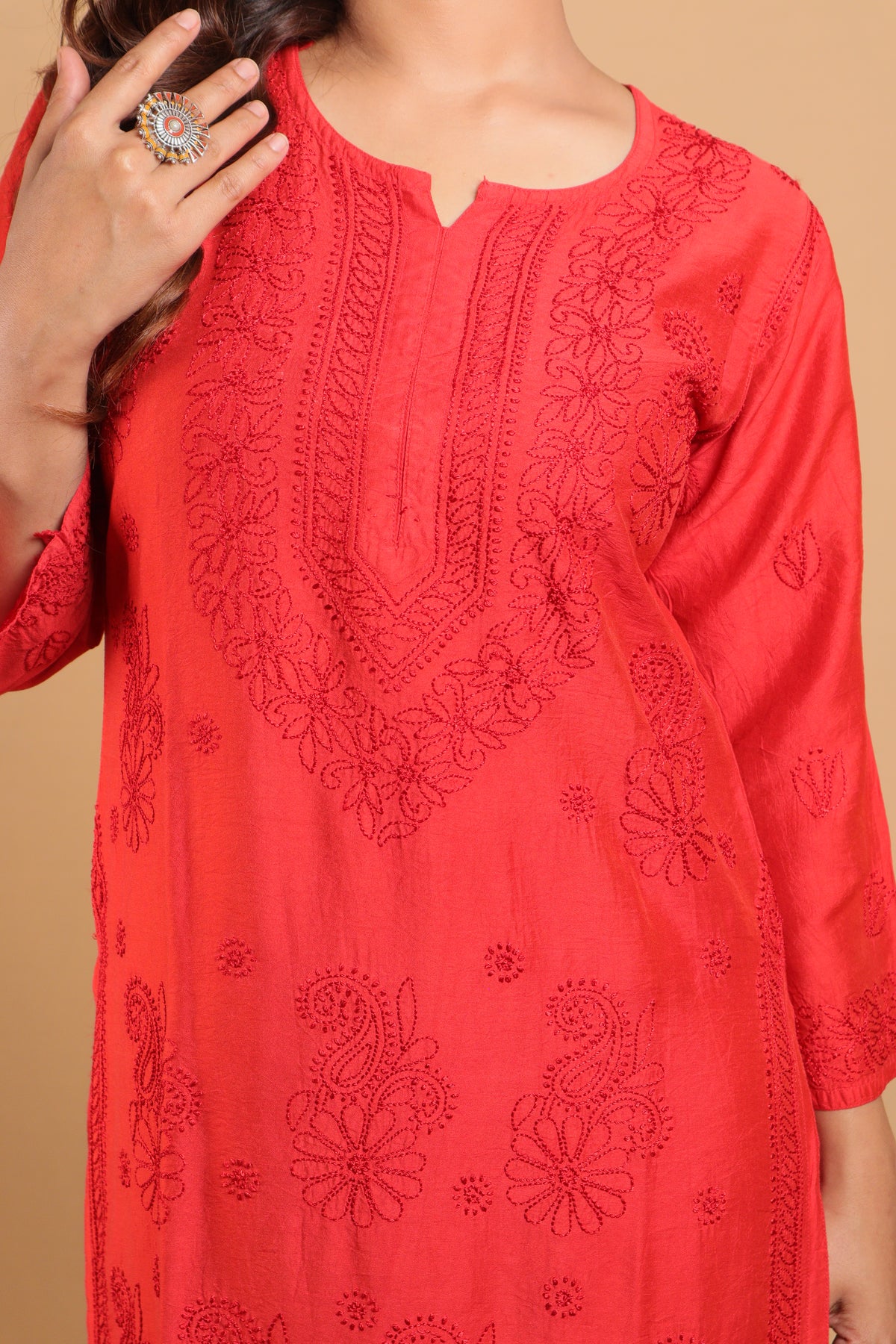 A close-up of a woman wearing a bright coral-red embroidered tunic top with floral and paisley cutwork details, set against a solid beige background.