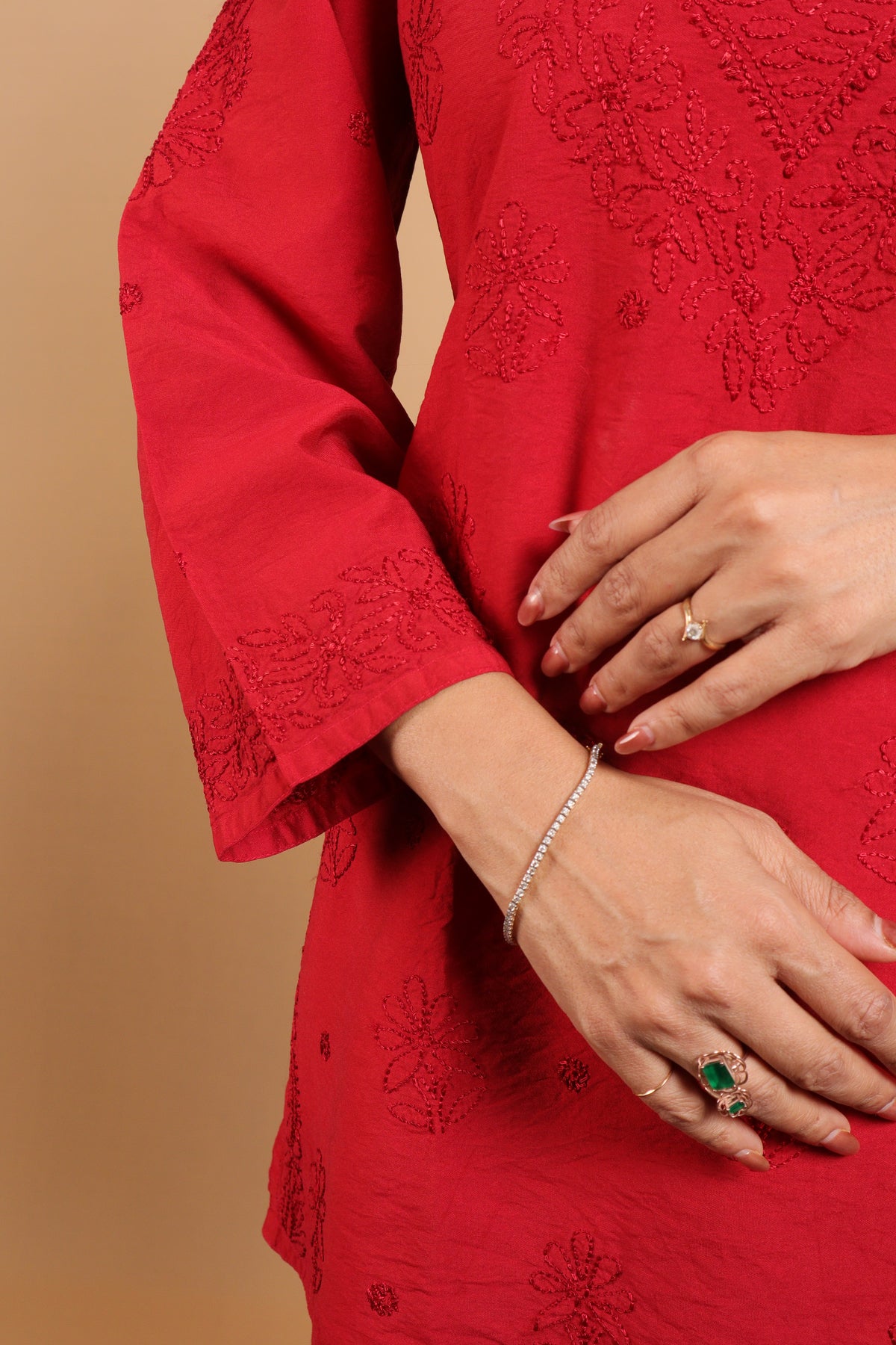 A close-up of a person wearing a bright red embroidered kurta or dress, with both hands visible against a warm beige studio background. The person is also wearing silver jewelry, including a bracelet and rings.