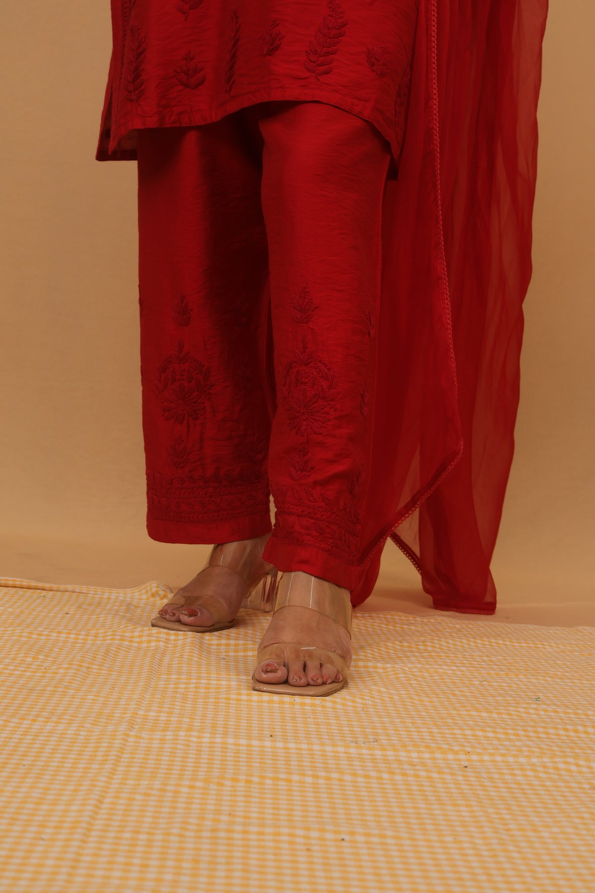 A person wearing a red traditional outfit with matching embroidered pants and a sheer red drape, paired with clear high-heeled sandals, standing against a beige backdrop.