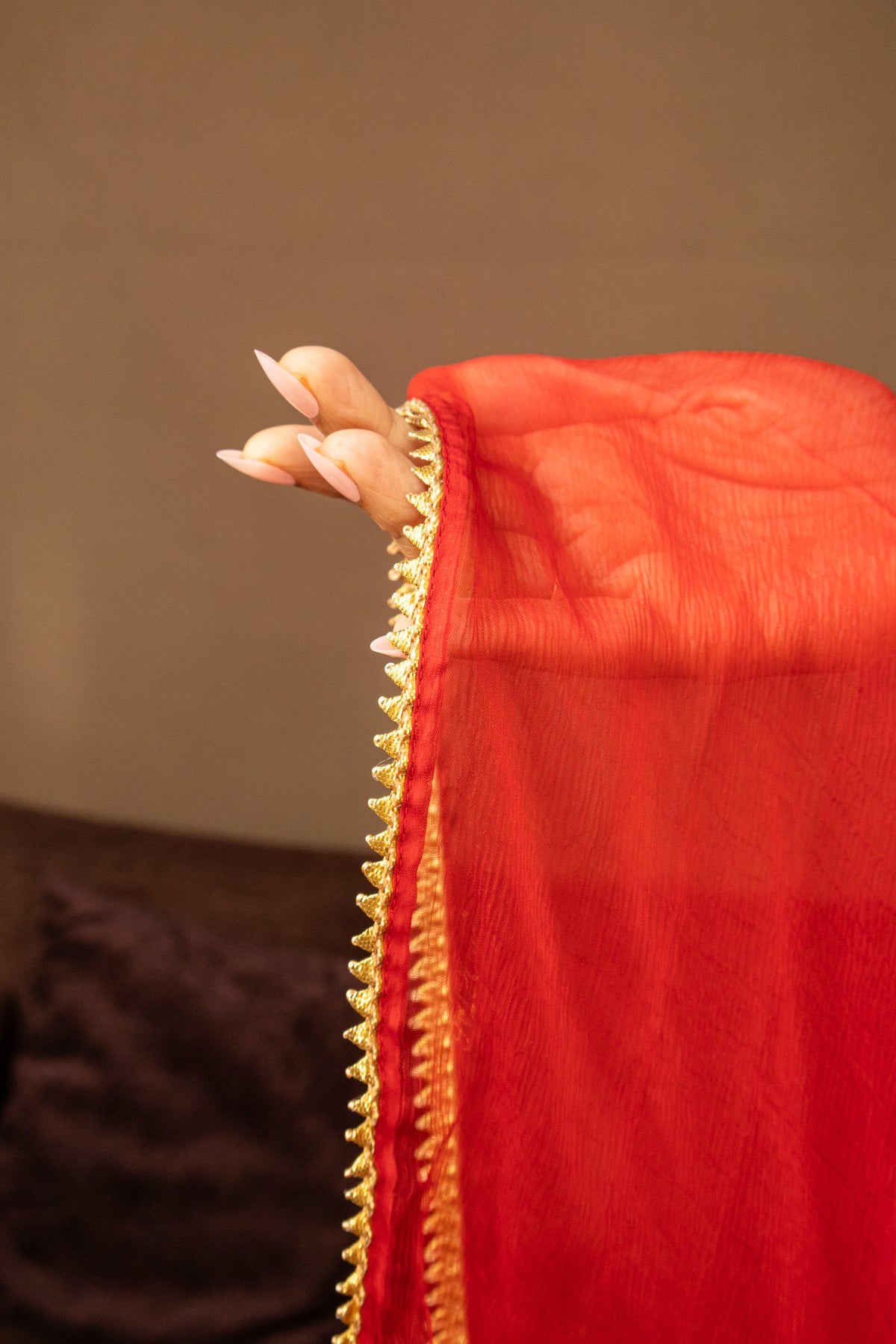 A close-up of a bright red dupatta or scarf draped over a person's hand, showing a decorative gold embroidered border.
