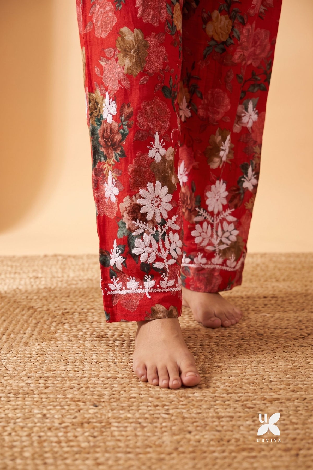 Red floral pajama pants with white and pink floral patterns and a white embroidered border, shown on a model's legs standing on a woven jute rug.