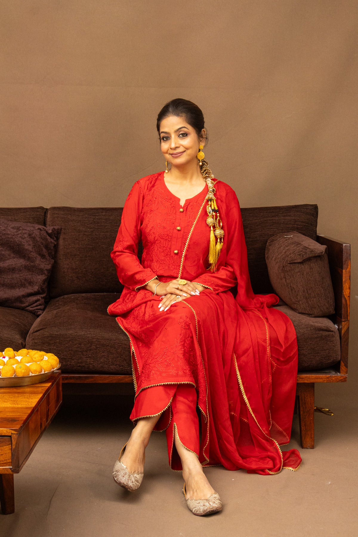 A woman wearing a traditional red Indian salwar kameez suit with gold zari embroidery sits on a brown sofa. The outfit includes a long-sleeved tunic, matching trousers, and a draped dupatta.
