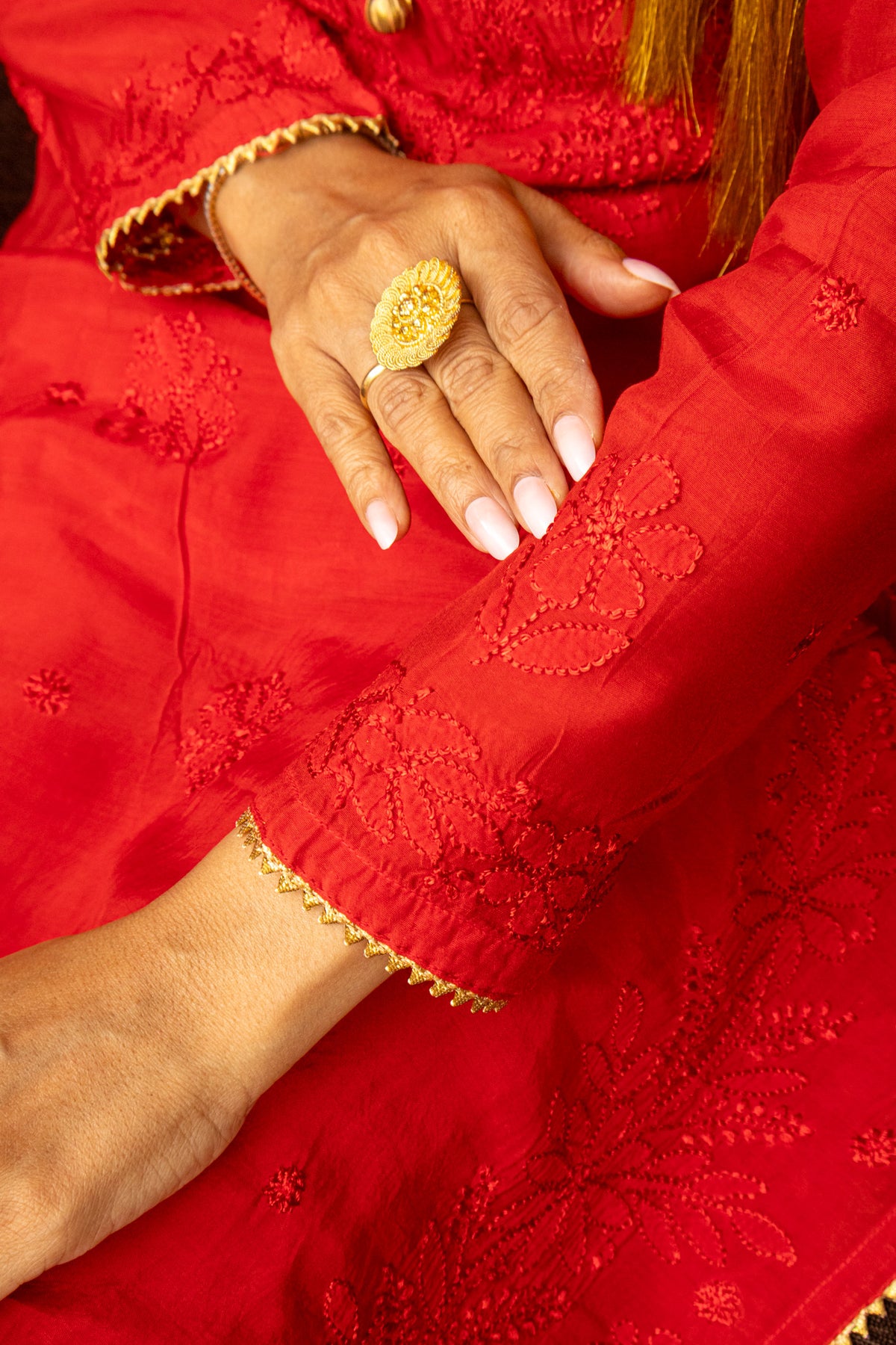 A close-up of a person's hand resting on a red embroidered garment, wearing a large gold ring with a floral design.