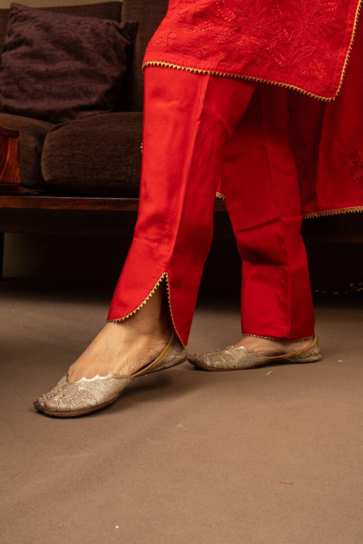 A person wearing bright red traditional pants with gold trim and matching red top, paired with metallic silver flat shoes, standing on a brown carpet in front of a dark sofa. Red Muslin Chikankari Bakhiya 2 Piece Set with Golden Gotta Lace