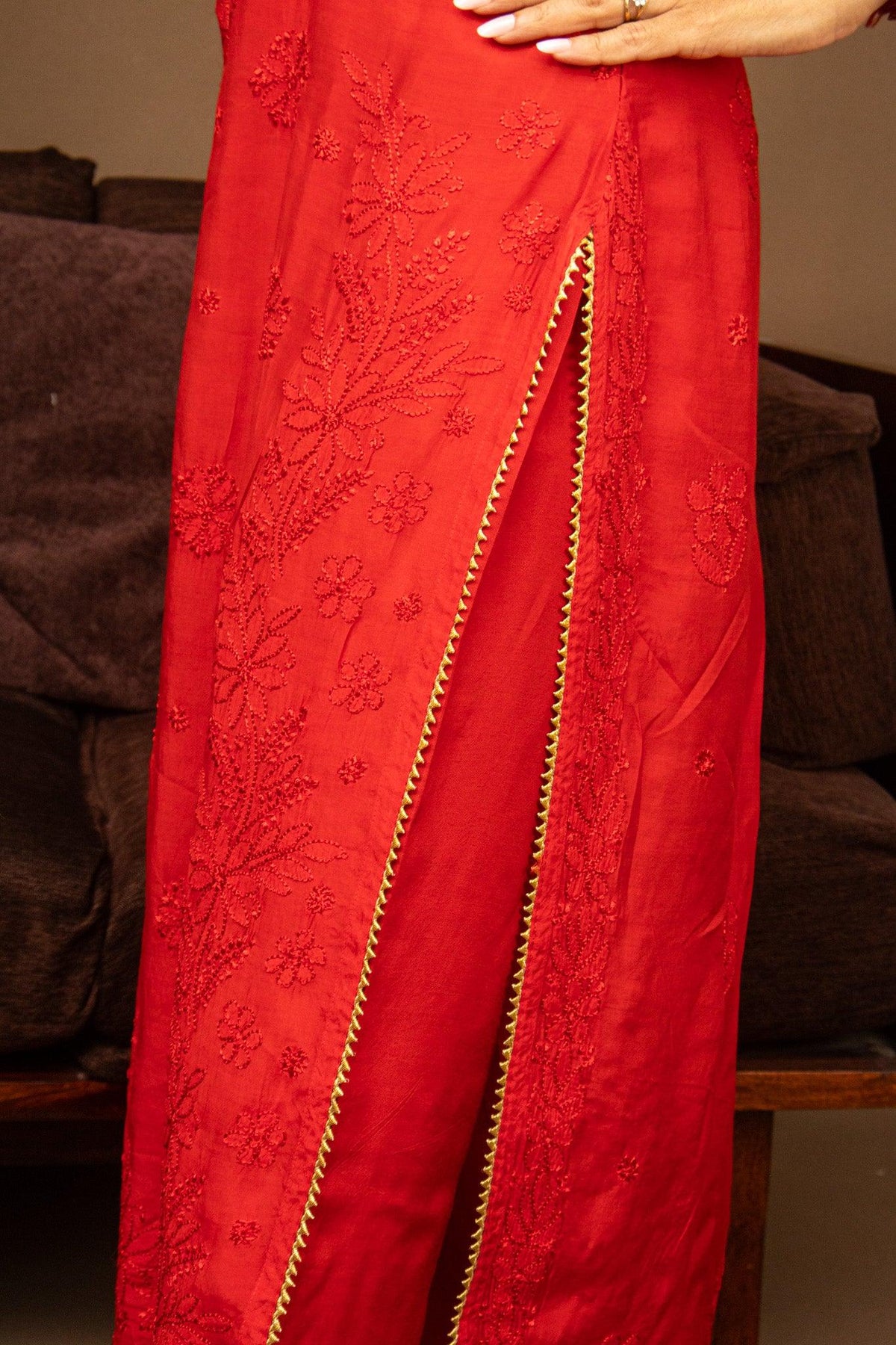 A close-up of a red embroidered dress or skirt with a side slit, featuring floral embroidery and a gold braided trim, worn by a model whose hand is visible at the top. Red Muslin Chikankari Bakhiya 2 Piece Set with Golden Gotta Lace
