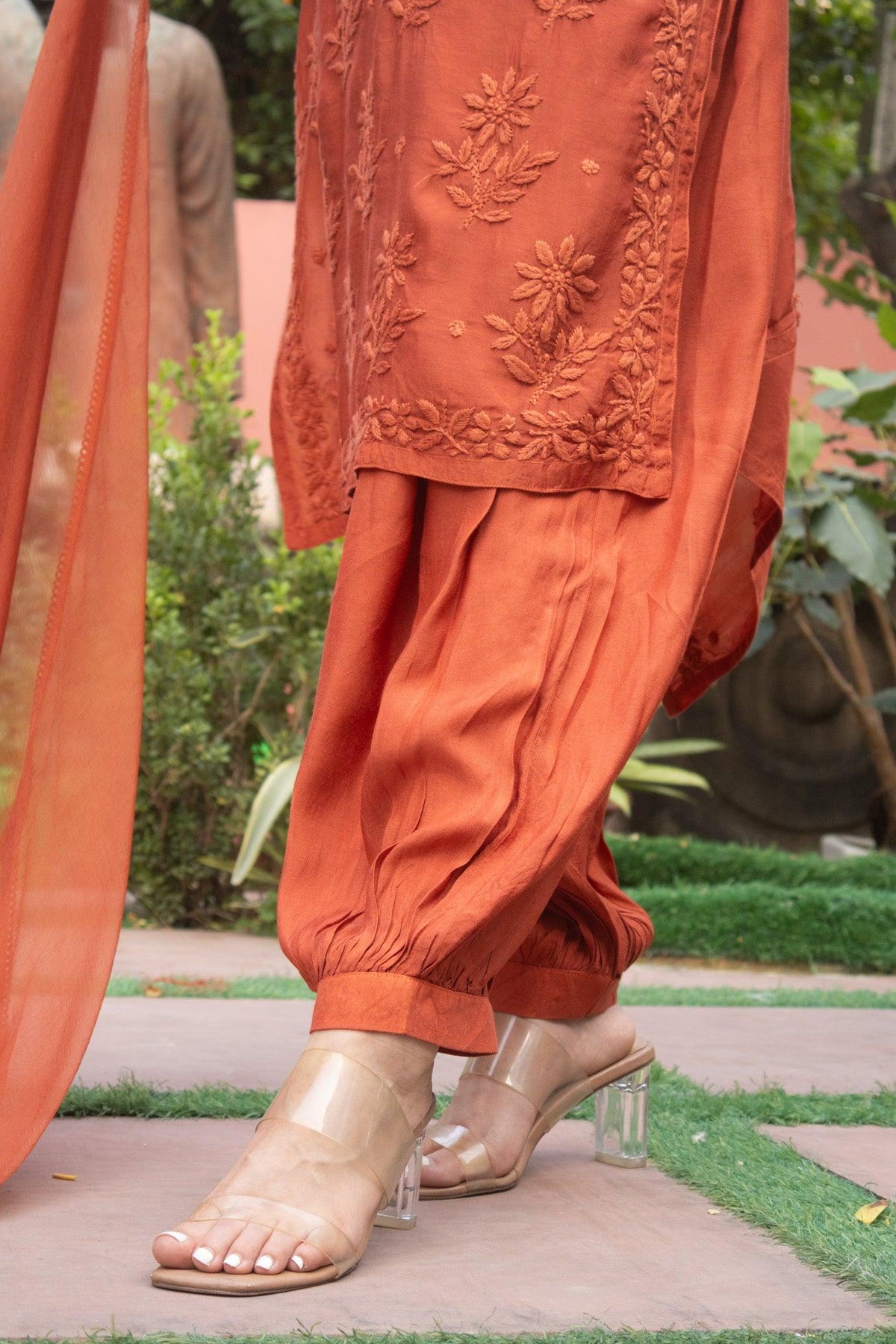 Close-up of a person wearing an orange rust-colored traditional outfit with matching embroidered kurta and draped pants, paired with clear high-heel sandals in an outdoor garden setting. Rust Red Muslin Chikankari Ghaaspatti Long Kurti with Tone-on-Tone Embroidery