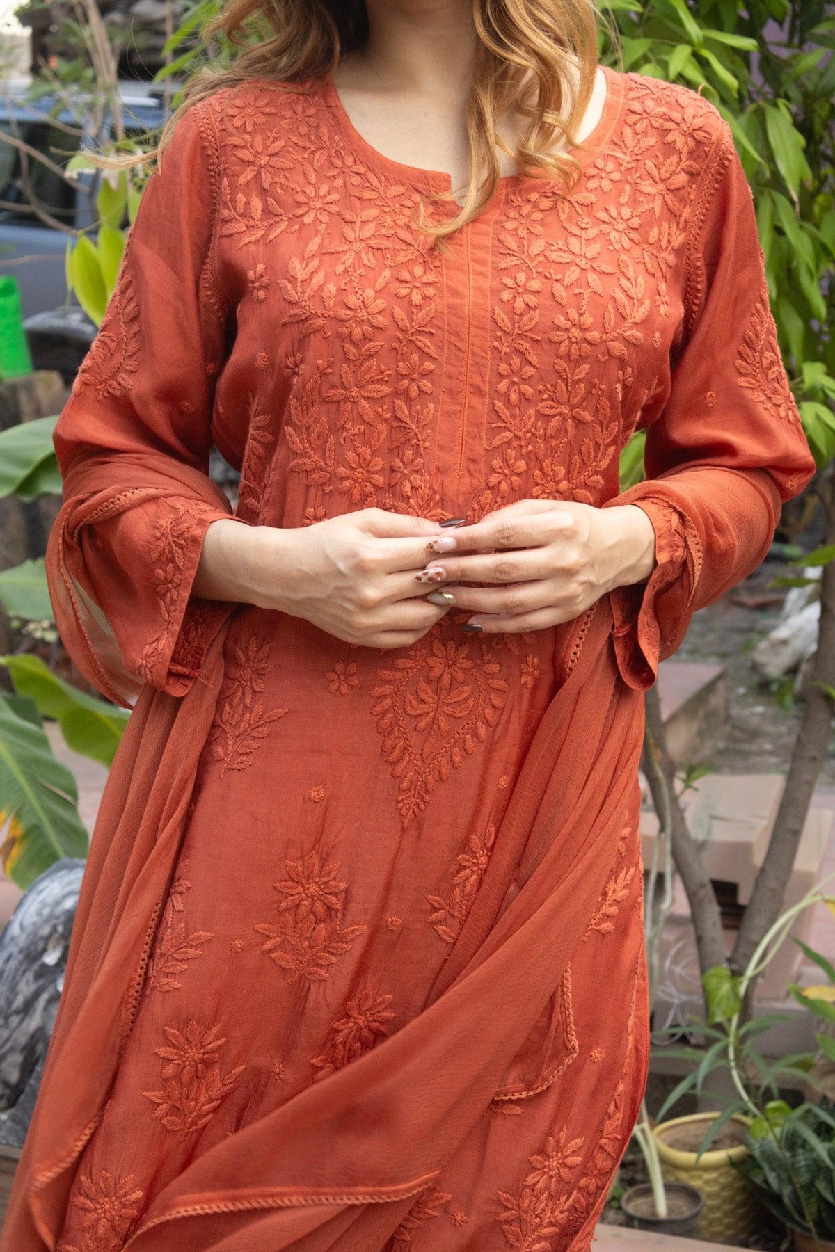 A woman wearing a rust-orange embroidered kurta with floral lace detailing, shown from the shoulders to the knees against an outdoor garden background. Rust Red Muslin Chikankari Ghaaspatti Long Kurti with Tone-on-Tone Embroidery