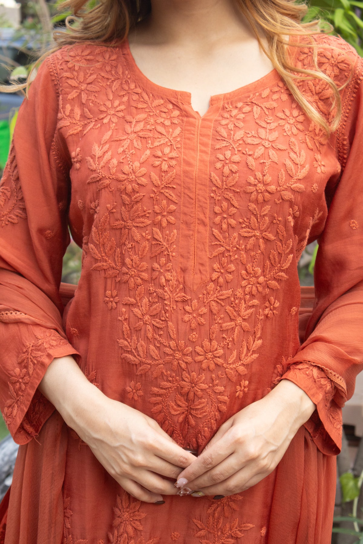 A close-up of a woman wearing a terracotta orange embroidered tunic with floral lace detailing, shown from the chest down in an outdoor setting.
