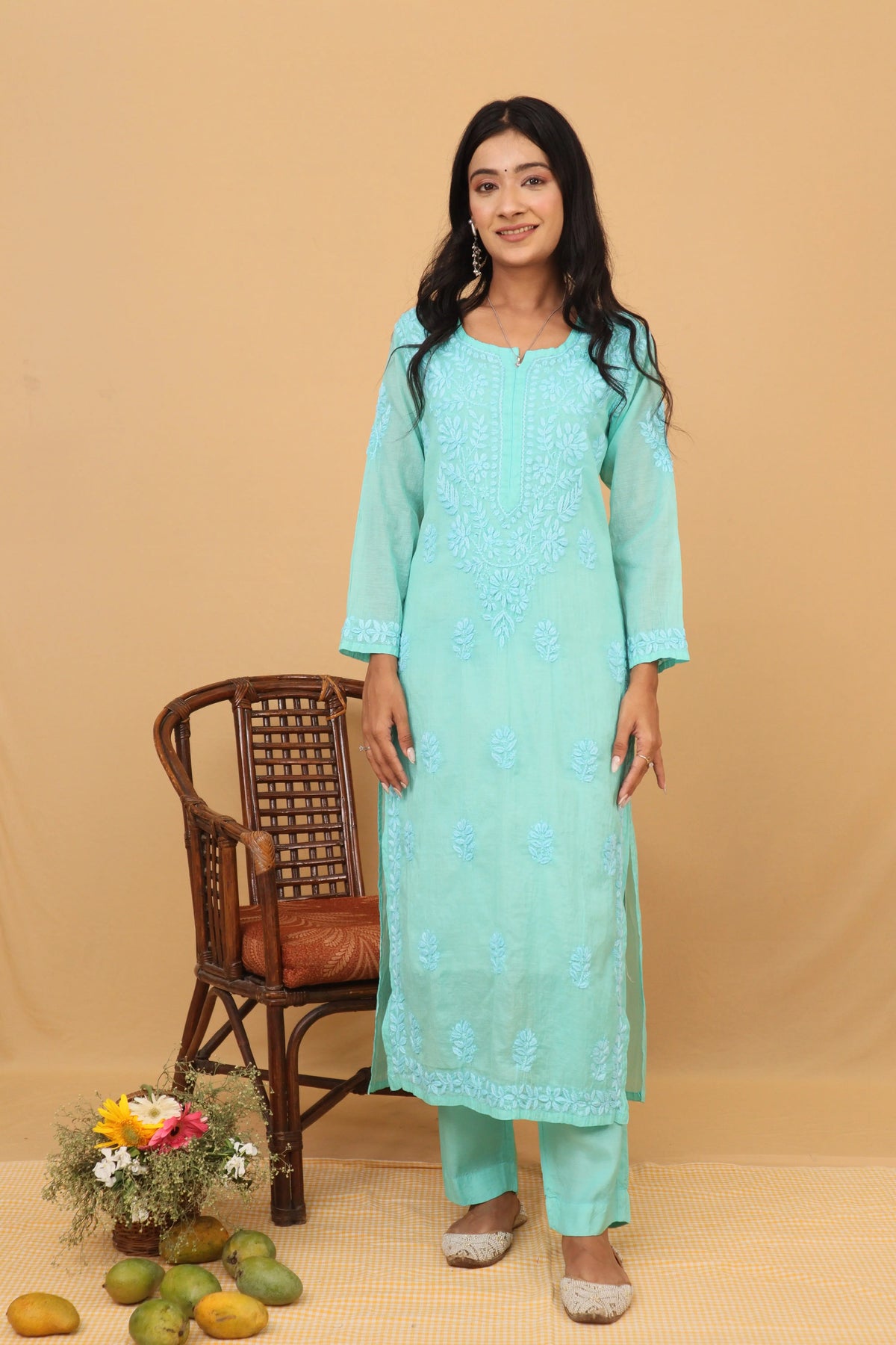 A woman modeling a mint green traditional Indian salwar kameez suit with white floral embroidery, standing next to a brown wicker chair on a beige studio background.