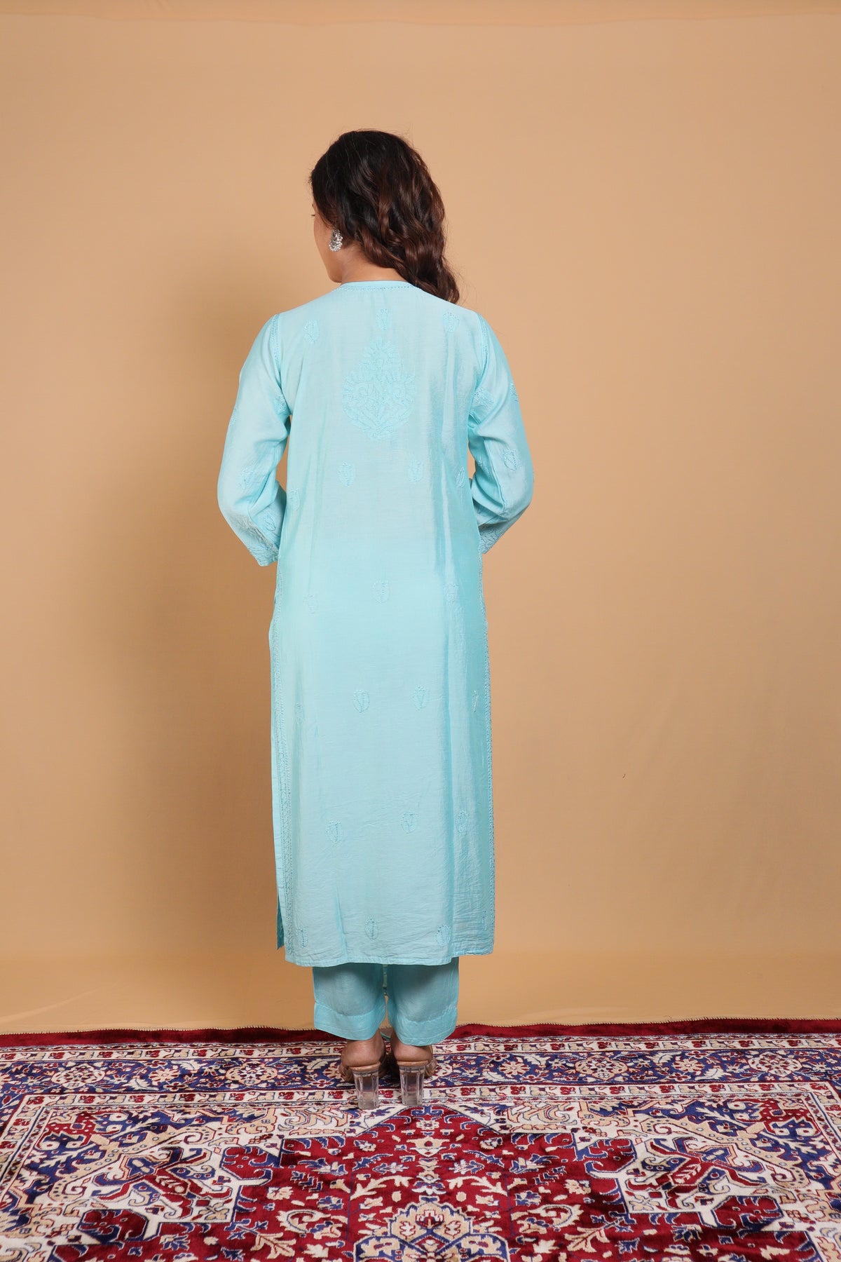 A woman viewed from behind wearing a light blue long kurta and matching pants, standing on a patterned red and blue rug against a solid beige backdrop.