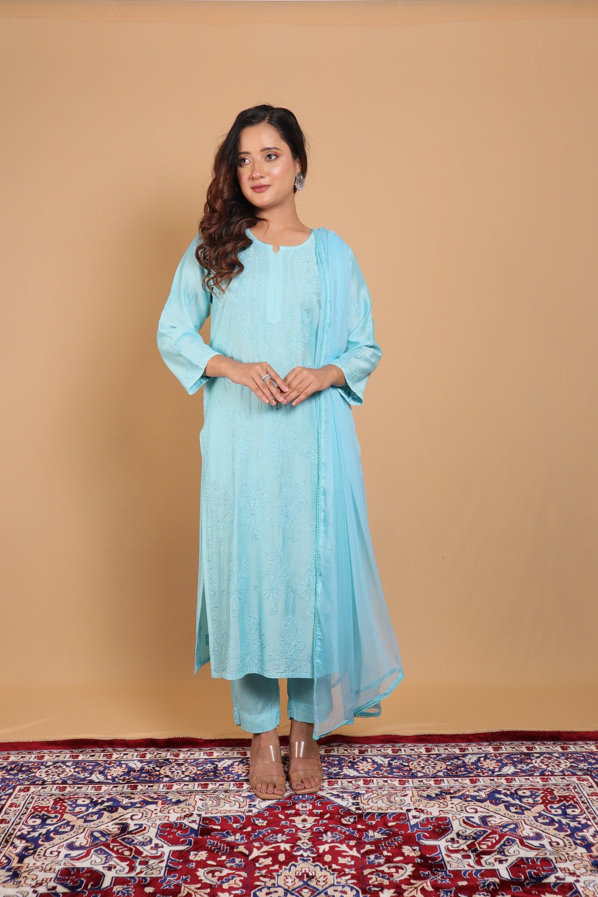 A woman modeling a light blue traditional outfit consisting of a long kurta, matching pants, and a sheer dupatta, standing on a patterned red and blue rug against a solid beige backdrop.