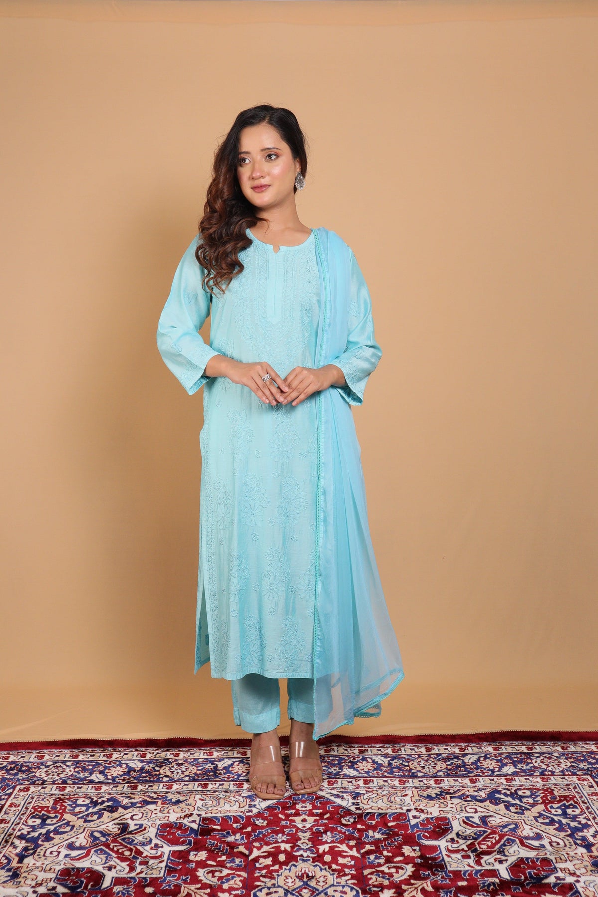 A woman modeling a light blue traditional outfit consisting of a long kurta, matching pants, and a sheer dupatta, standing on a patterned red and blue rug against a solid beige backdrop.
