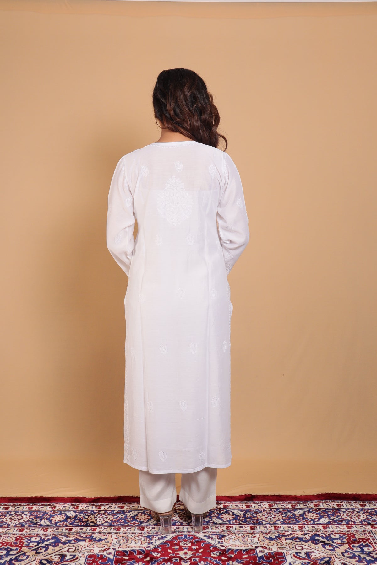 A woman viewed from behind wearing a long white tunic and matching pants, standing on a patterned rug against a plain beige backdrop.