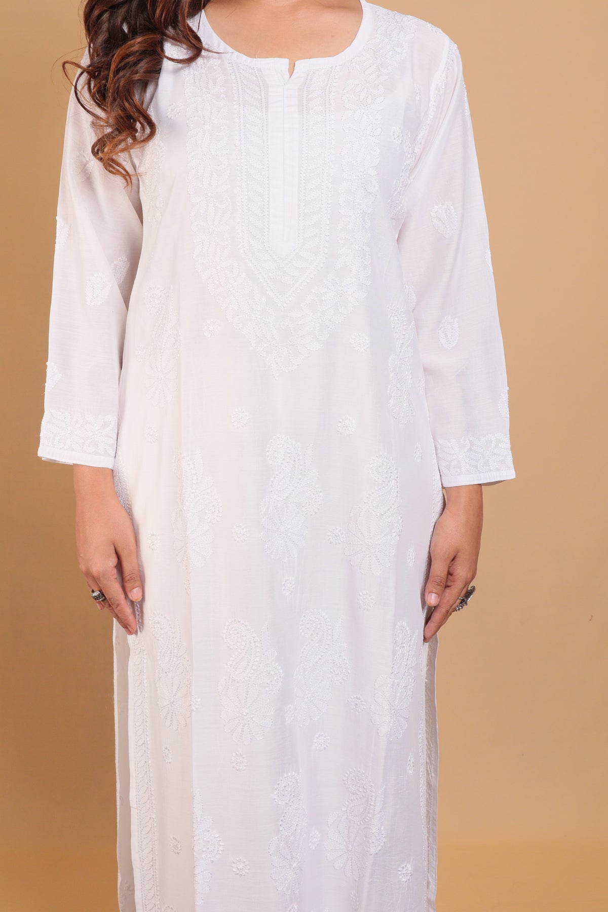 A woman wearing a white long kurta with delicate floral embroidery, shown from the neck down against a solid beige background.