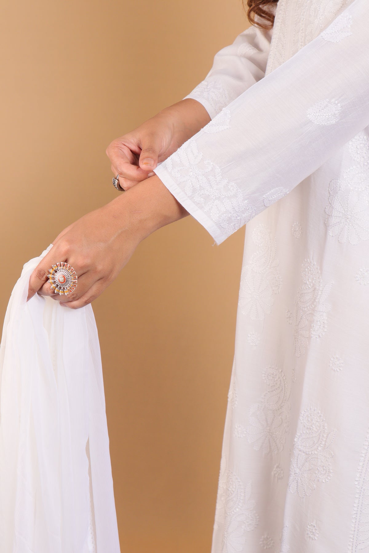 A close-up of a person wearing a white embroidered traditional outfit with a matching sheer white dupatta, holding it against a warm beige background.