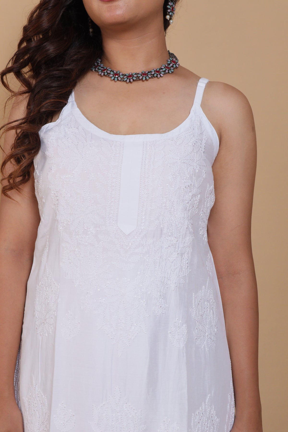 A woman wearing a white sleeveless camisole dress with delicate lace embroidery, paired with a blue beaded choker necklace, set against a solid beige background.