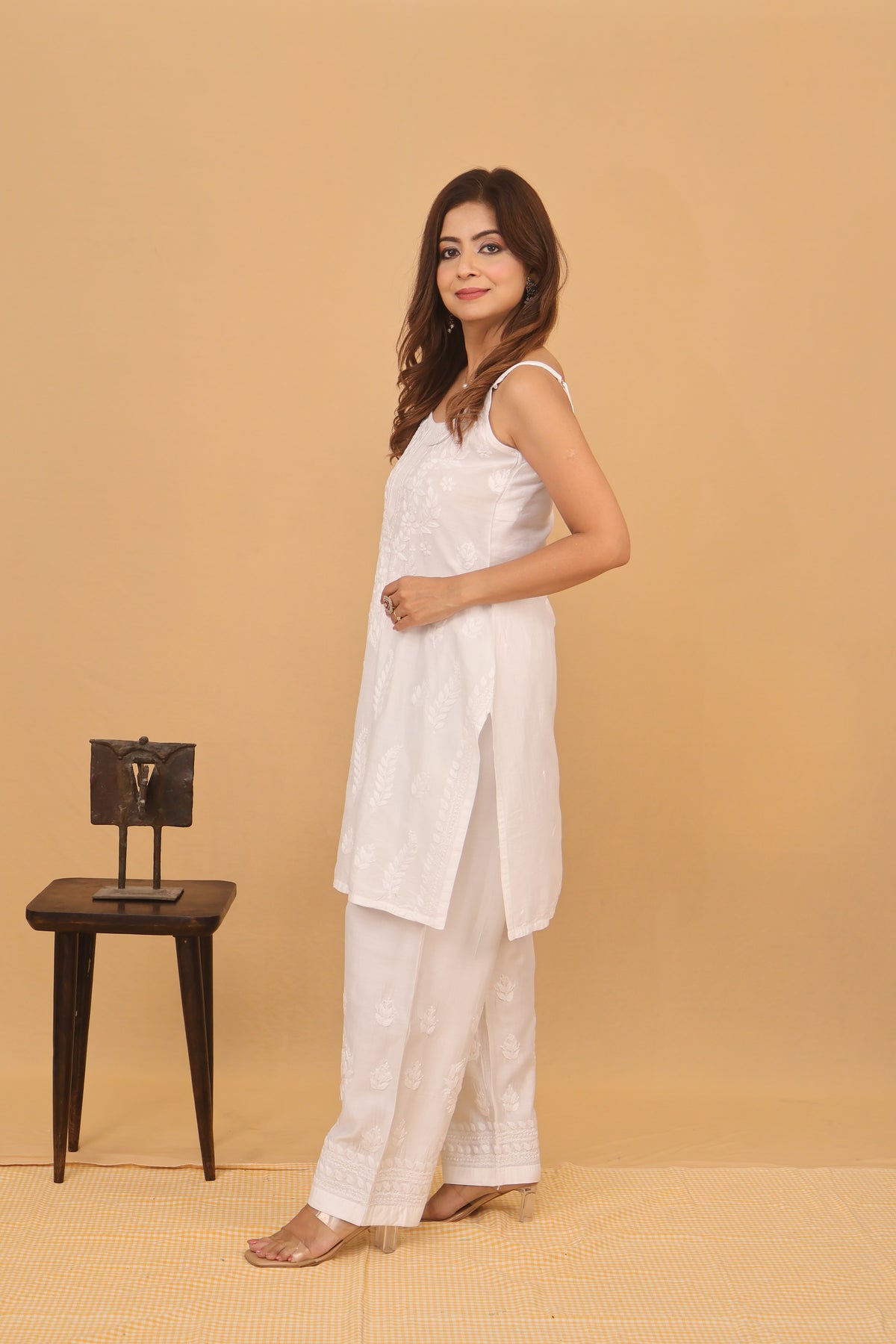 A woman modeling a white embroidered kurta and wide-leg pants set against a solid beige studio backdrop, standing next to a small dark wooden stand.