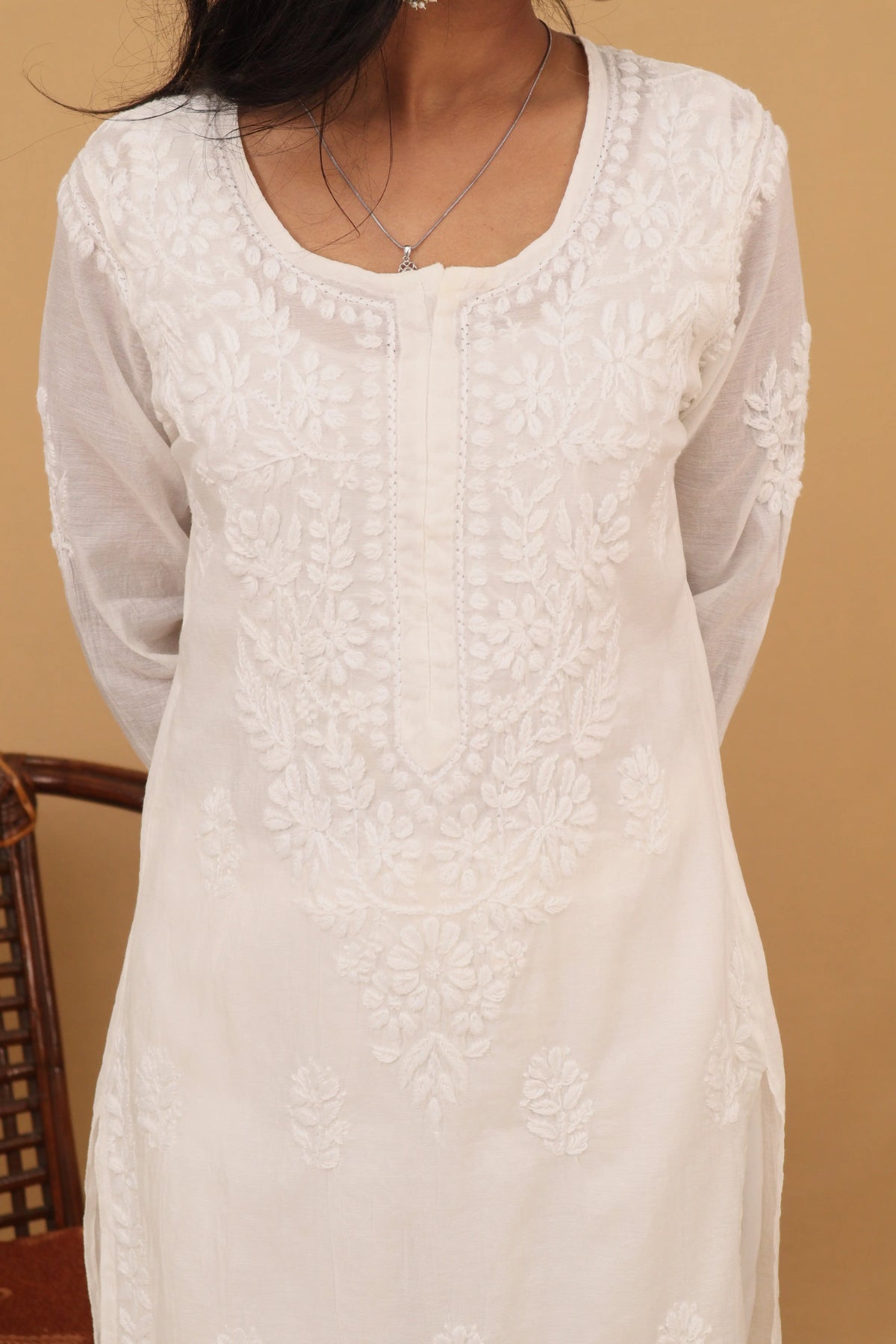 A woman wearing a cream-colored embroidered kurta with floral lace detailing on the bodice and sleeves, set against a warm beige background.