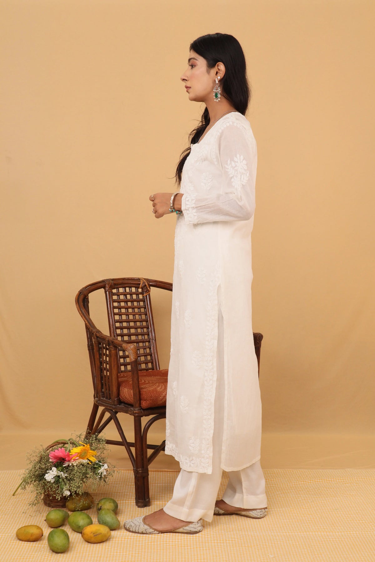 A woman stands in profile wearing a long white embroidered kurta with matching white pants, posed next to a brown wicker chair on a beige studio backdrop with decorative fruit and flowers on the floor.