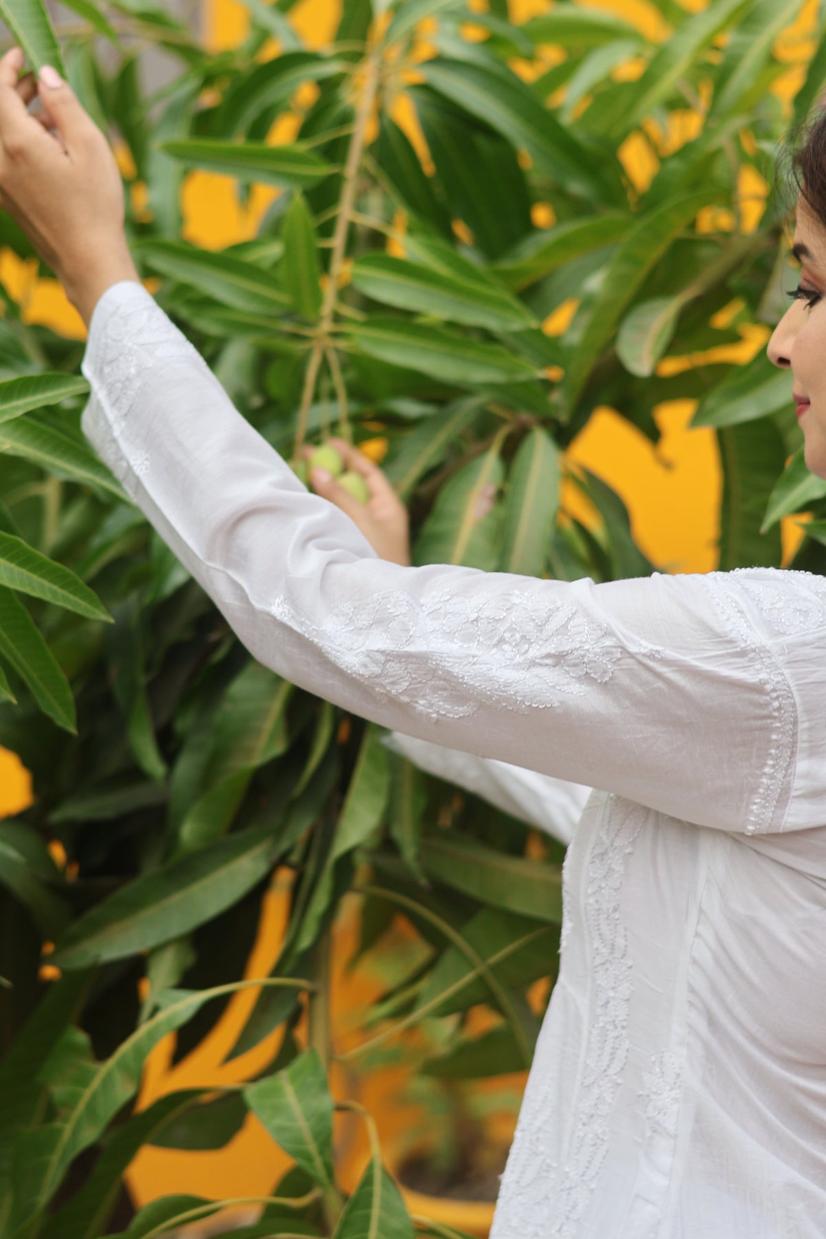 A woman in a white embroidered kurta reaches toward green leaves against a bright yellow background.