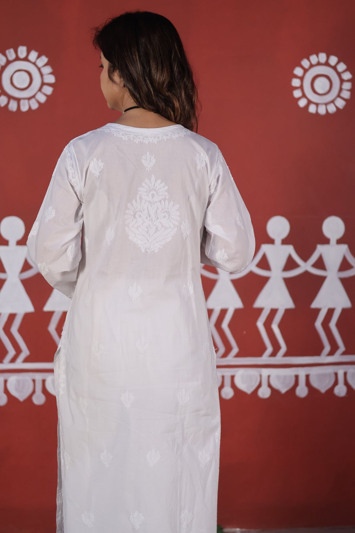 A woman viewed from behind wearing a white long-sleeve kurta with white printed motifs, standing against a red wall decorated with white folk art figures.