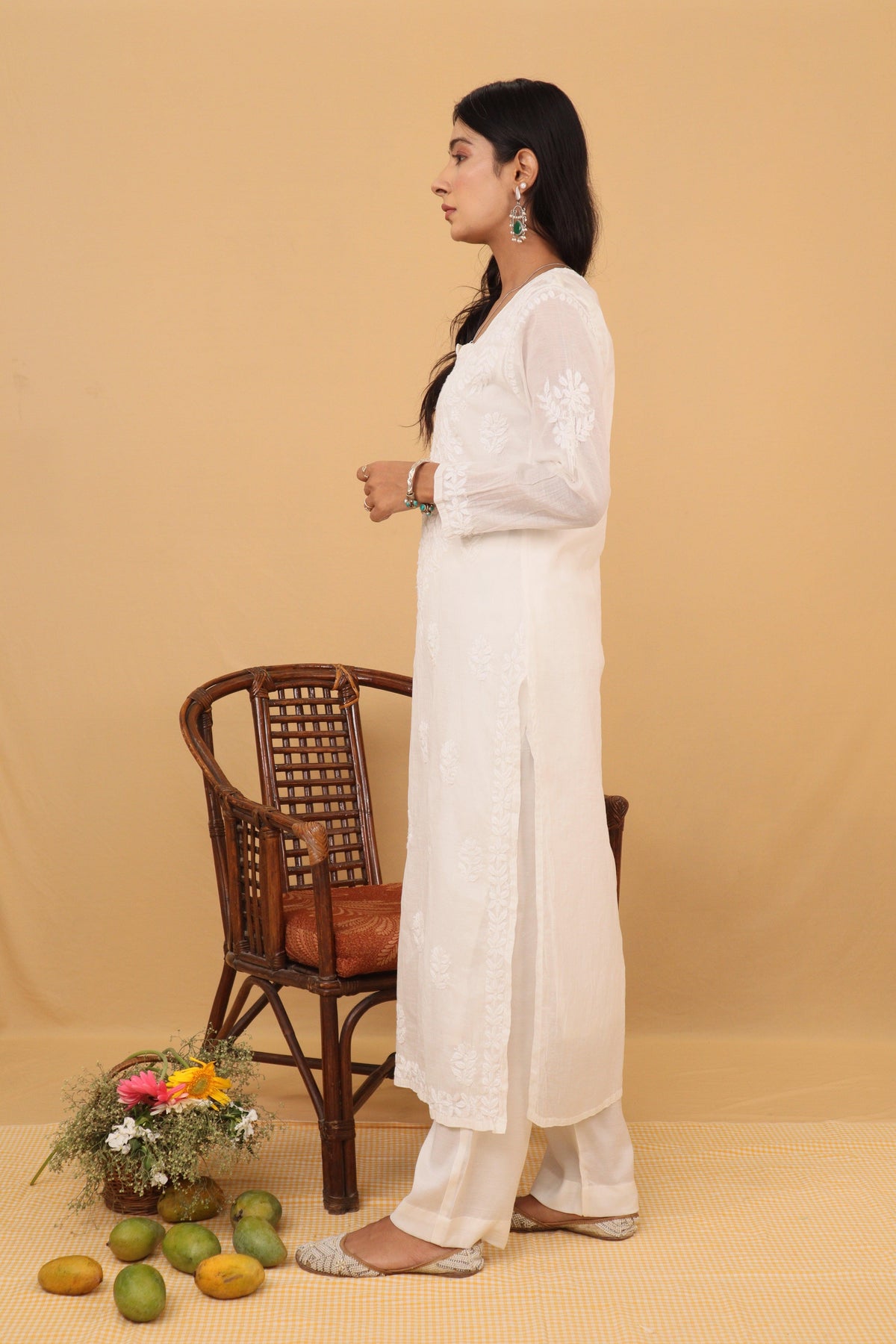 A woman stands in profile wearing a long white embroidered kurta with matching white pants against a warm beige studio backdrop. A brown woven chair, a small flower arrangement, and a few green fruits are placed on the floor nearby. White Mul Chanderi Ghaspatti Chikankari Long Kurti 2 Piece Set