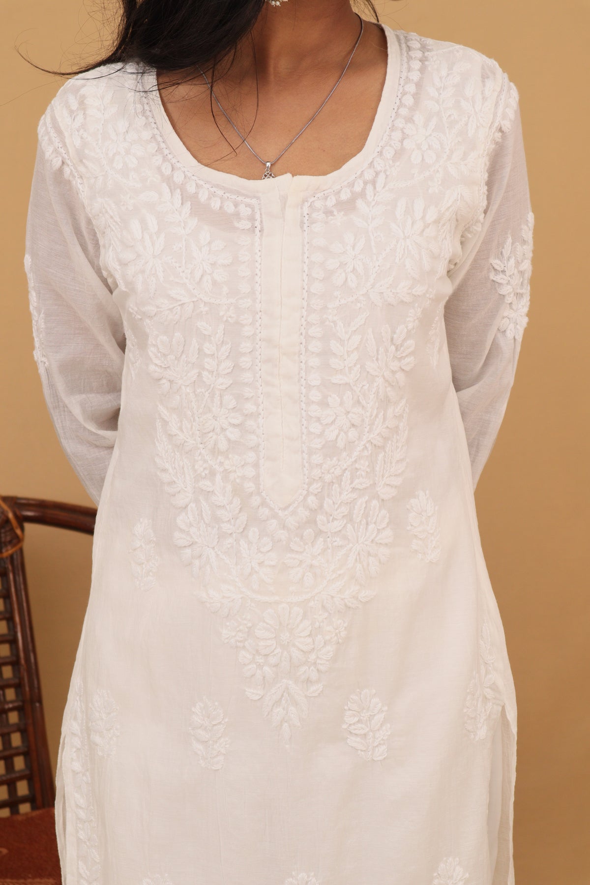 A woman wearing a white embroidered tunic top with floral lace detailing and sheer long sleeves, shown against a warm beige background.