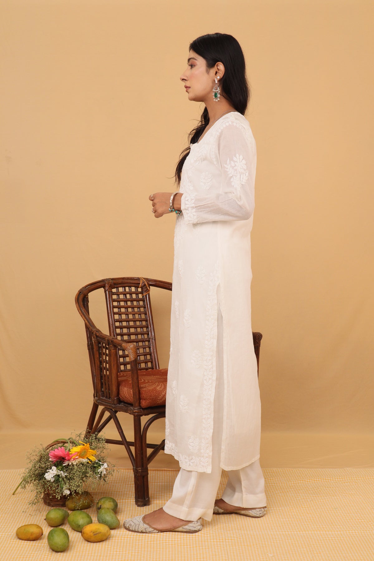 A woman stands in profile wearing a long white embroidered kurta with matching white pants against a warm beige studio backdrop. A brown woven chair, a small flower arrangement, and a few green fruits are placed on the floor nearby.