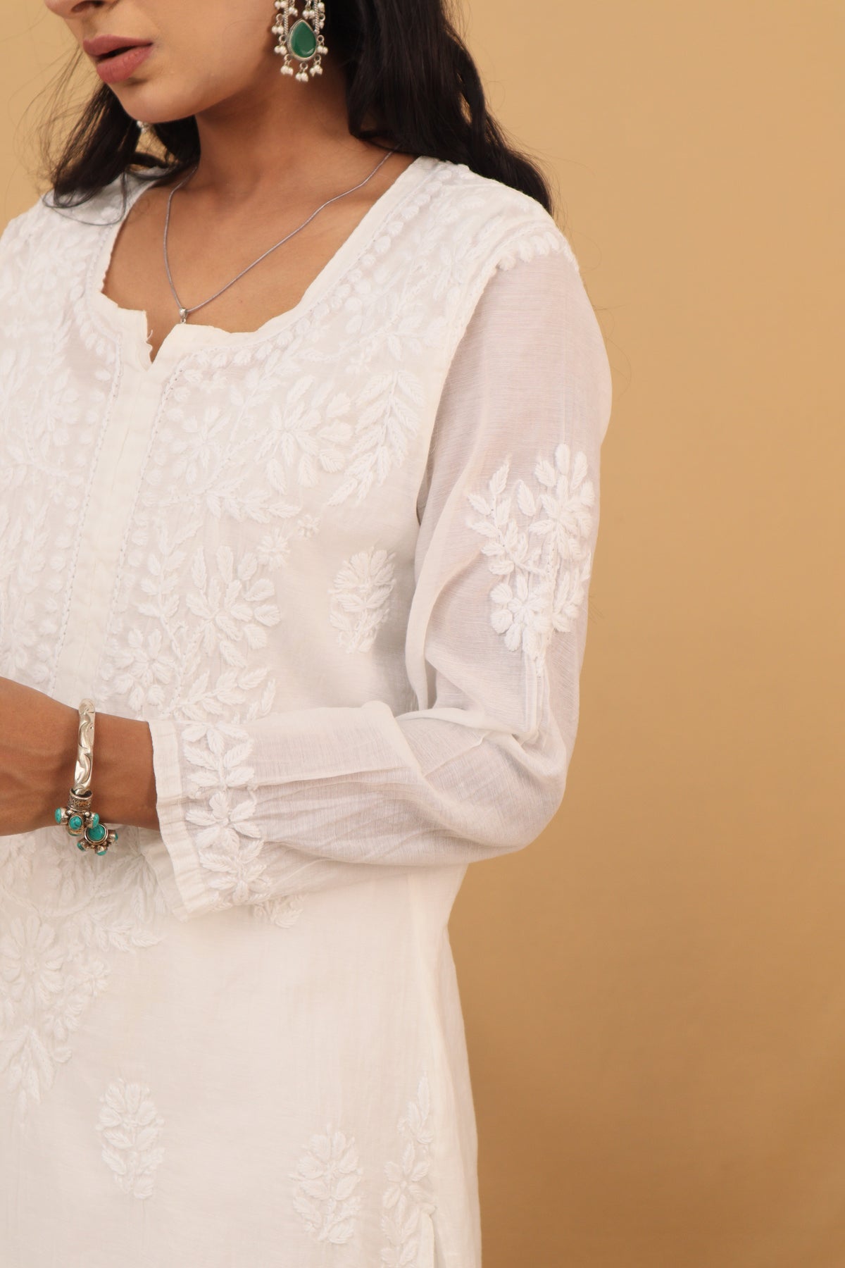 A close-up of a woman wearing a white embroidered kurta with floral lace detailing on the sleeves and bodice, paired with green-accented jewelry against a warm beige background.
