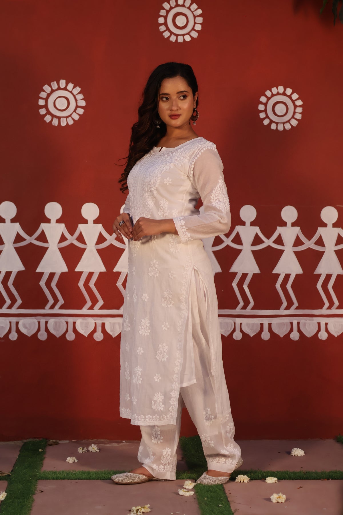 A woman modeling a white embroidered ethnic suit with a kurta, pants, and dupatta, standing in front of a red wall decorated with white folk art figures.