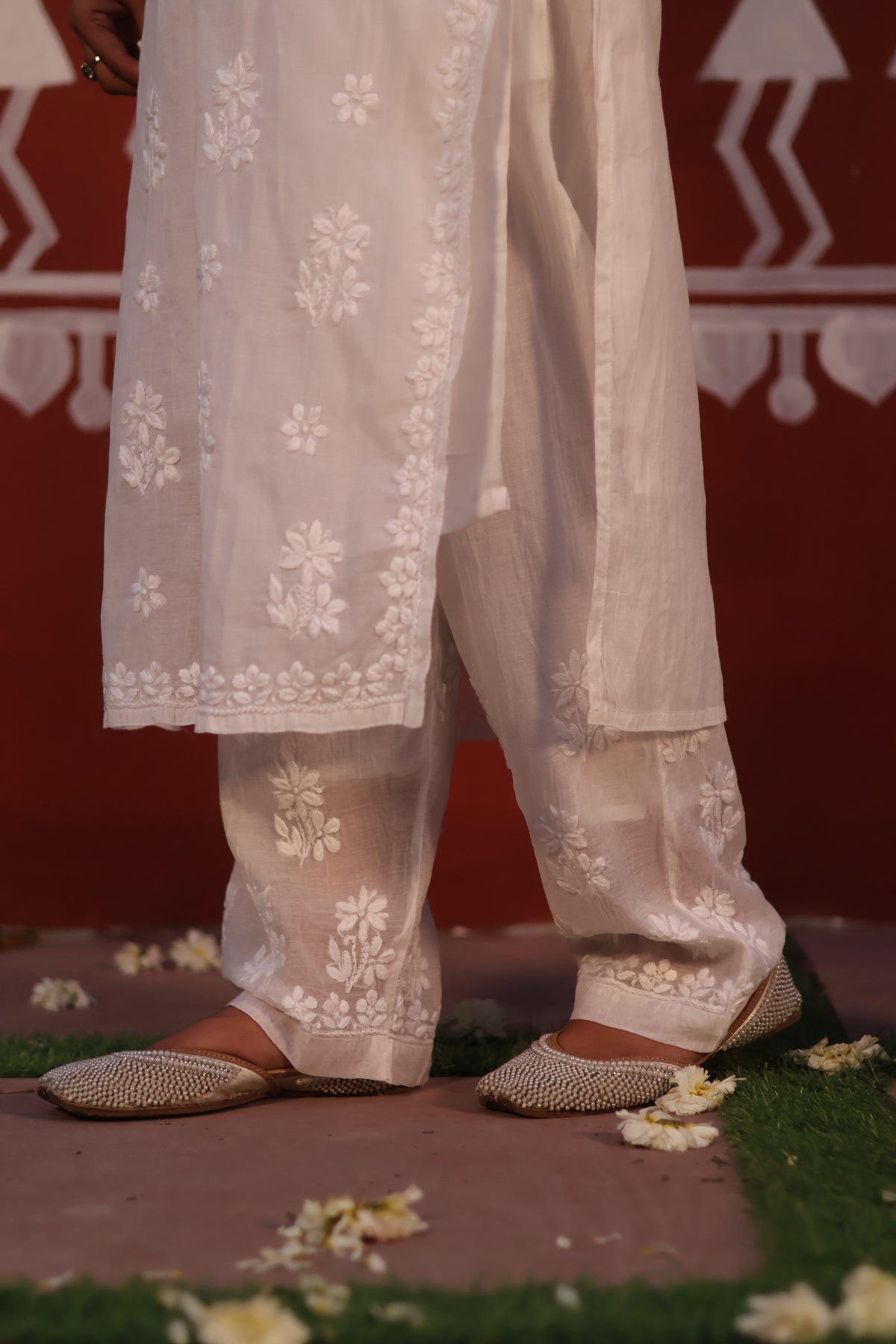 A close-up of a person wearing white embroidered traditional pants with matching flat shoes, standing on a decorative red and green set with flower petals scattered around.
