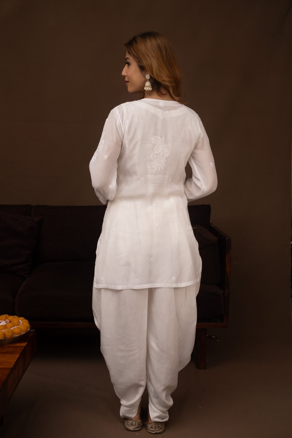 A woman shown from the back wearing a white traditional outfit with a long-sleeve top and loose pants, standing in a dimly lit indoor setting with a dark sofa behind her.