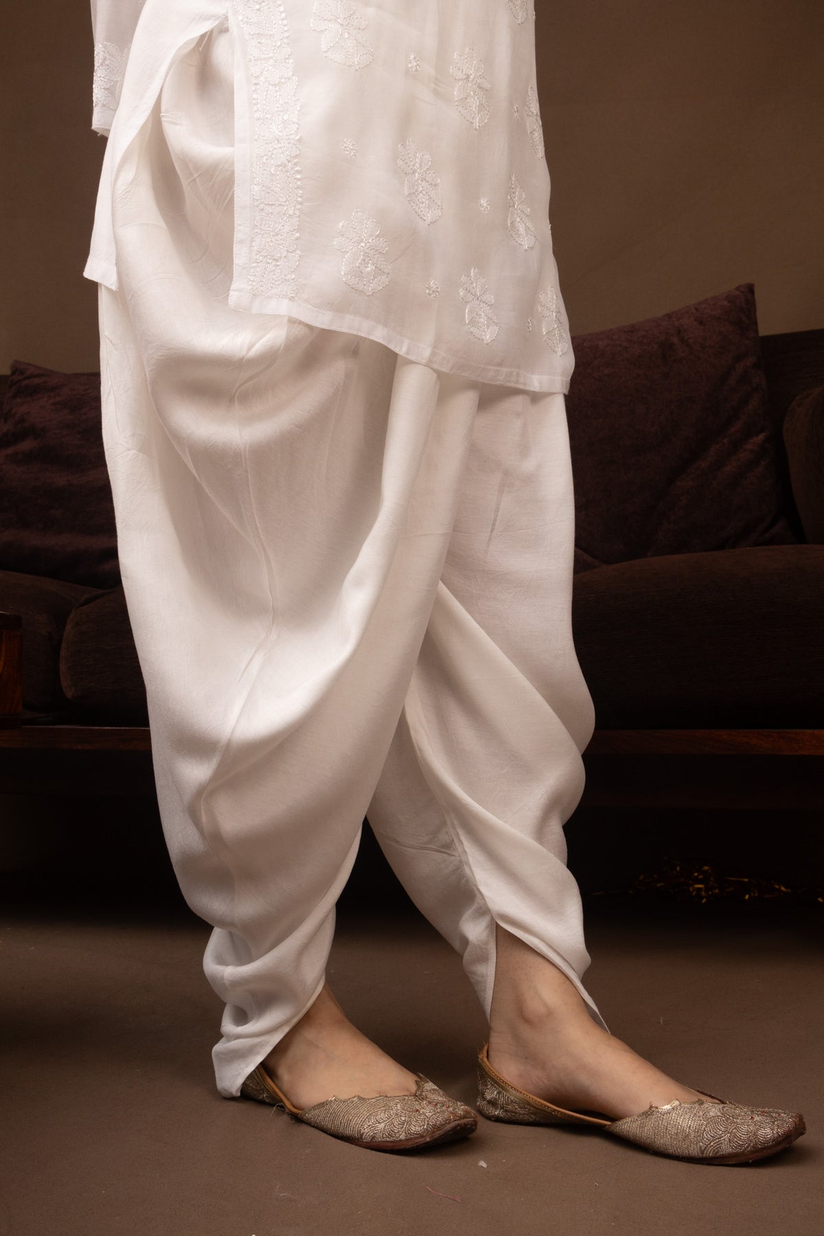 A person wearing loose white traditional draped pants with a matching embroidered white top and flat beige shoes, standing indoors in front of a dark brown couch.