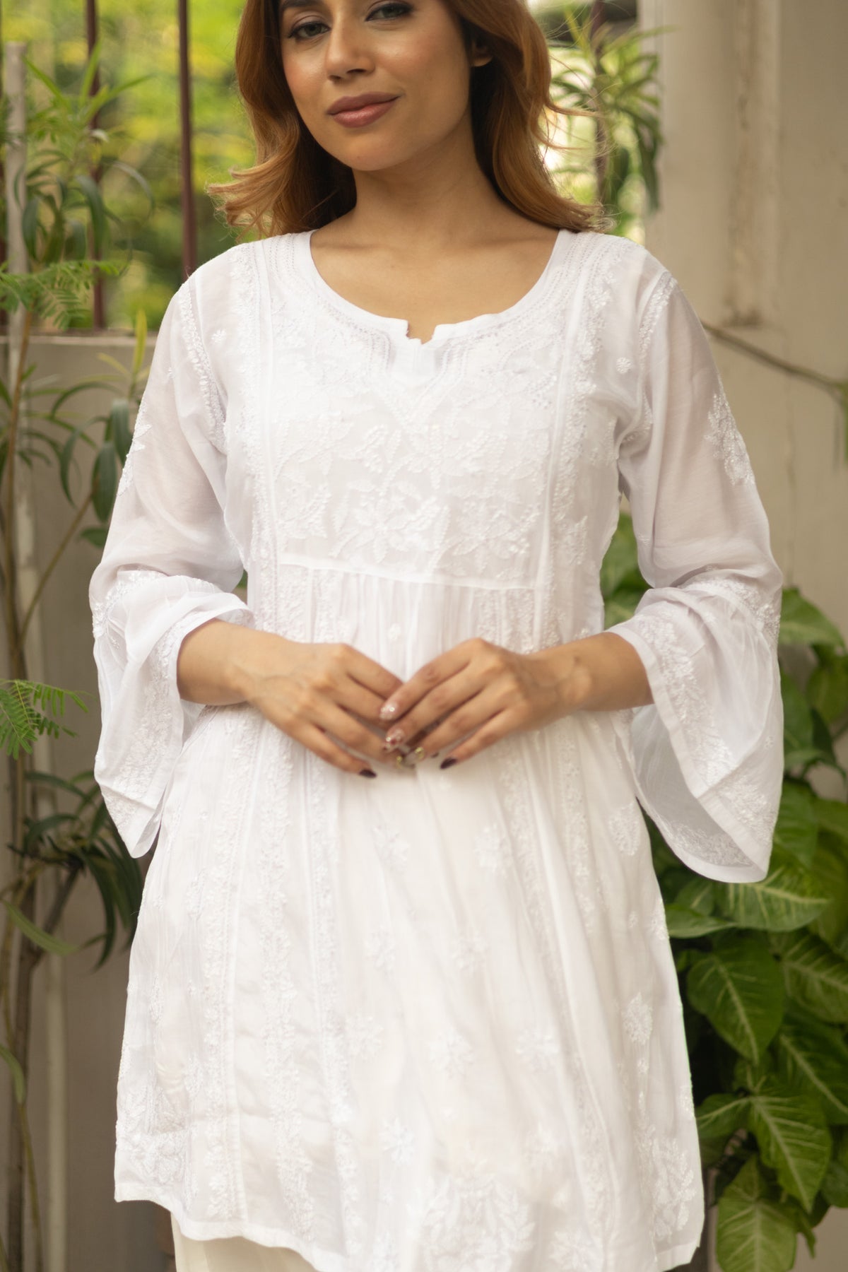 A woman wearing a white embroidered cotton tunic with three-quarter bell sleeves, standing in front of a background of green plants and a light-colored wall.