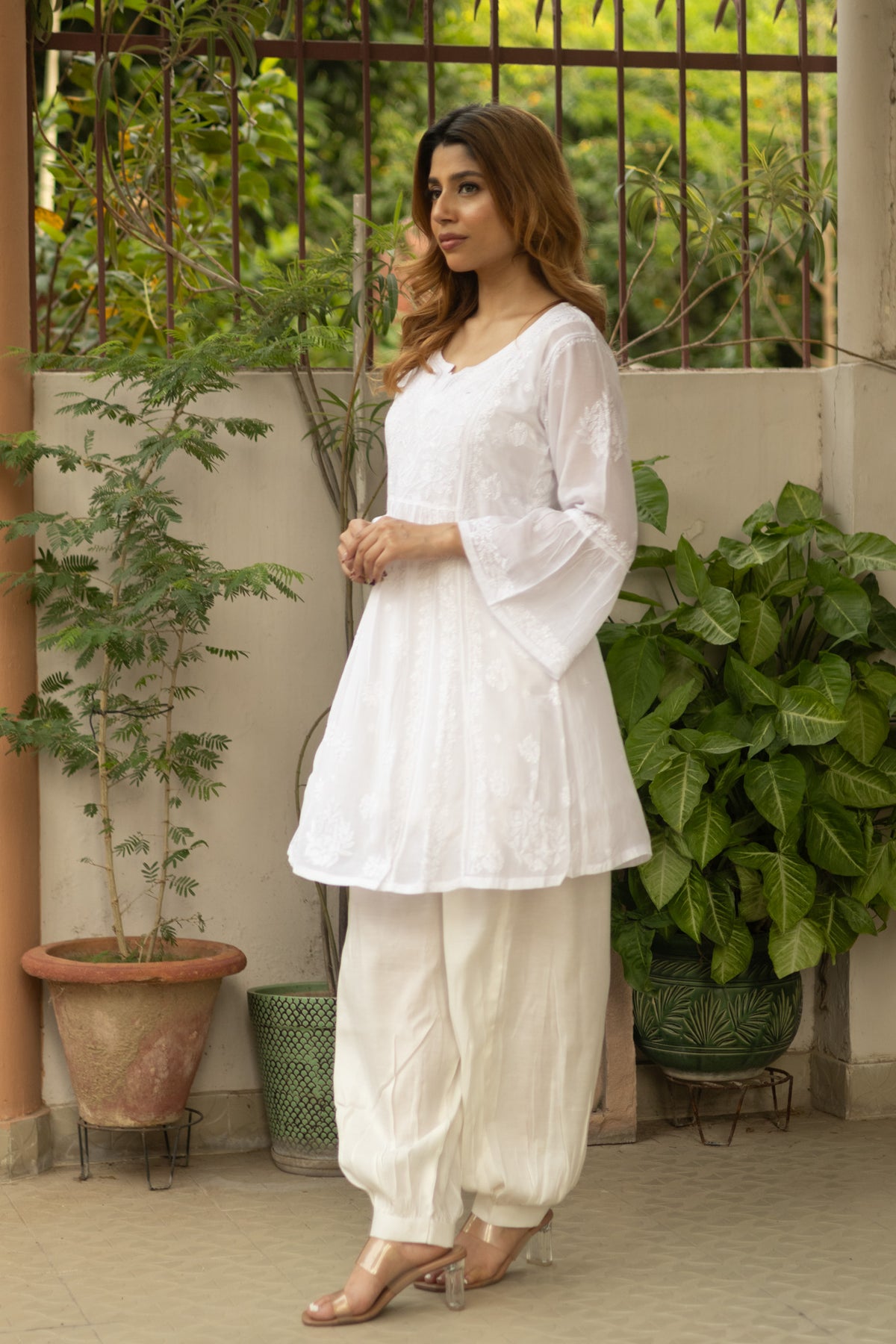 A woman modeling a white embroidered kurta with flared sleeves and matching white harem pants, standing in an outdoor patio setting with potted plants.