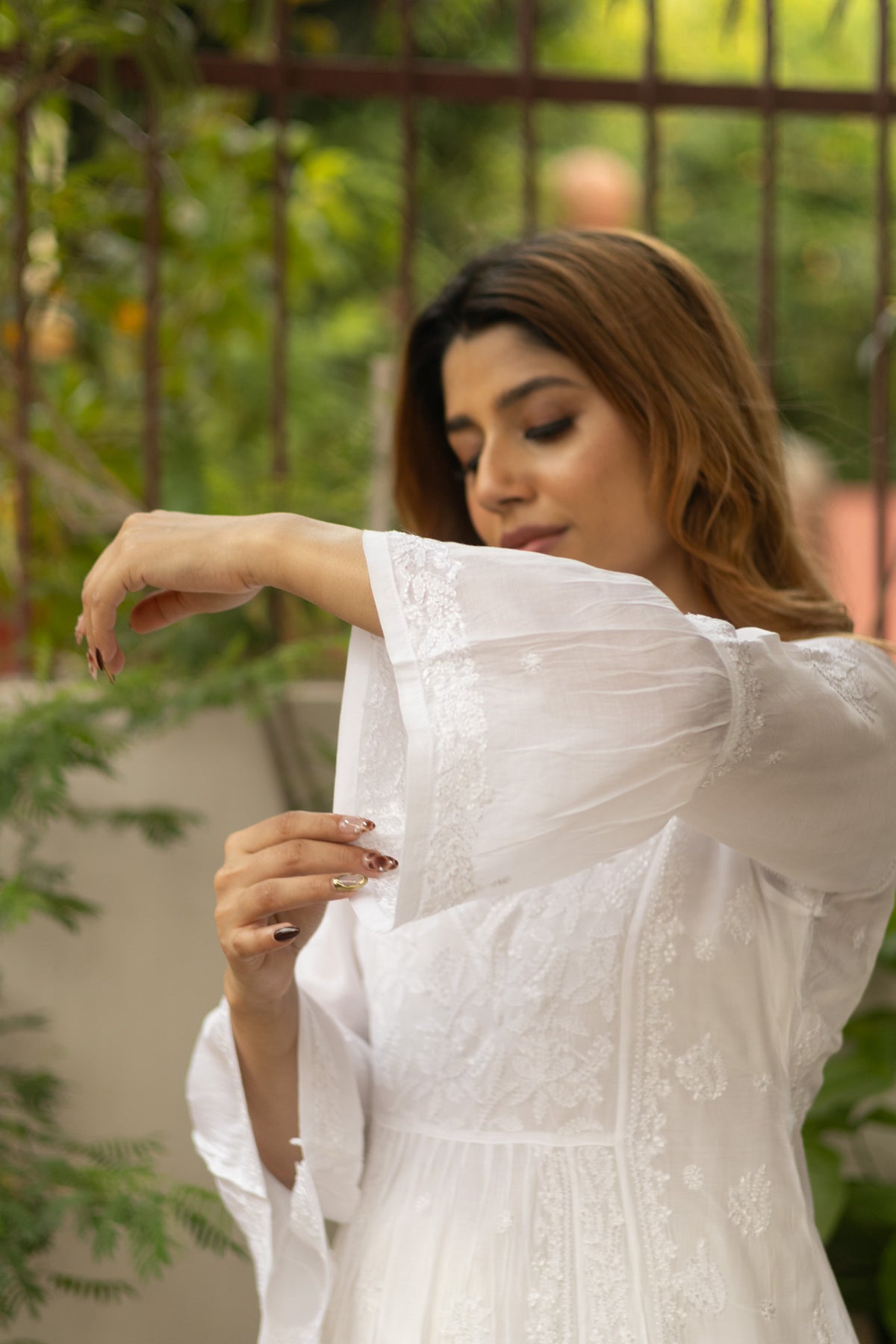 A woman wearing a white embroidered long-sleeve dress with sheer puff sleeves, shown in an outdoor setting with green foliage and a metal fence in the background.